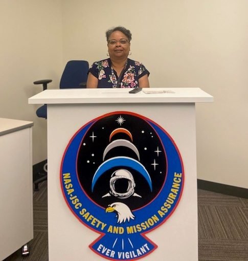 A woman in professional attire and wearing glasses sits behind a large, white podium with the seal of NASA-JSC Safety and Mission Assurance on the front.