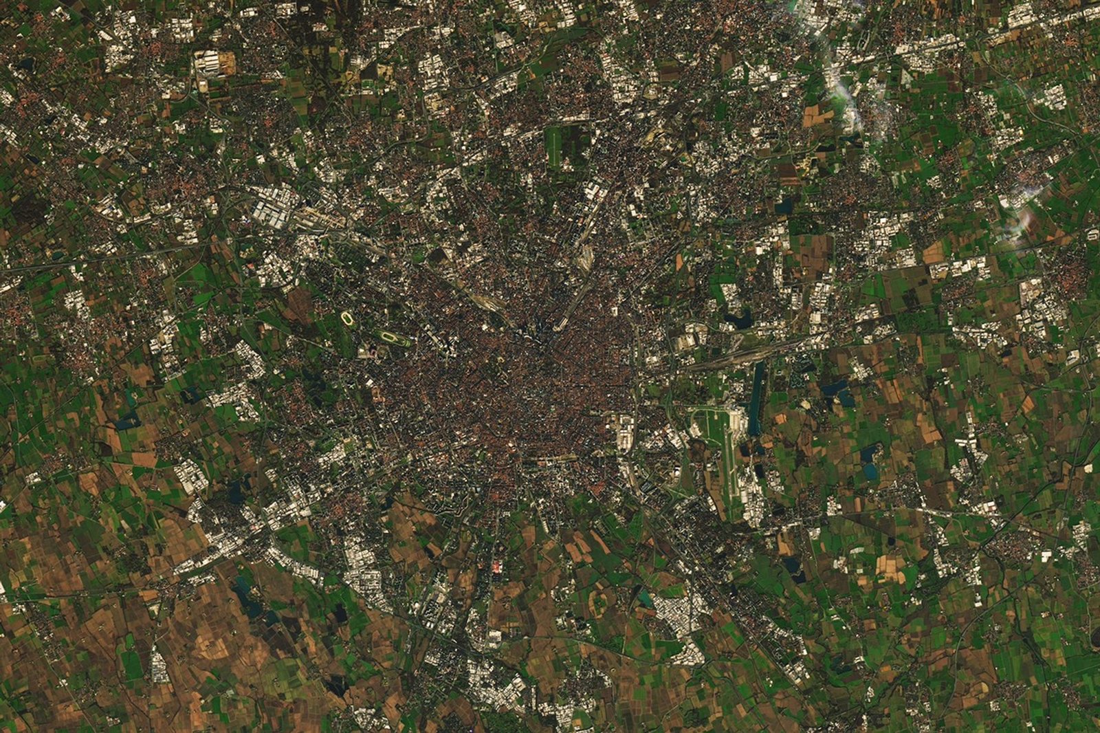 Satellite image of Milan, Italy, showing gray and dark-brown urban areas surrounded by green and tan agricultural land. Multiple Olympic sports venues are labeled and highlighted with white boxes, including ice hockey and speed skating locations, each with inset detailed views.