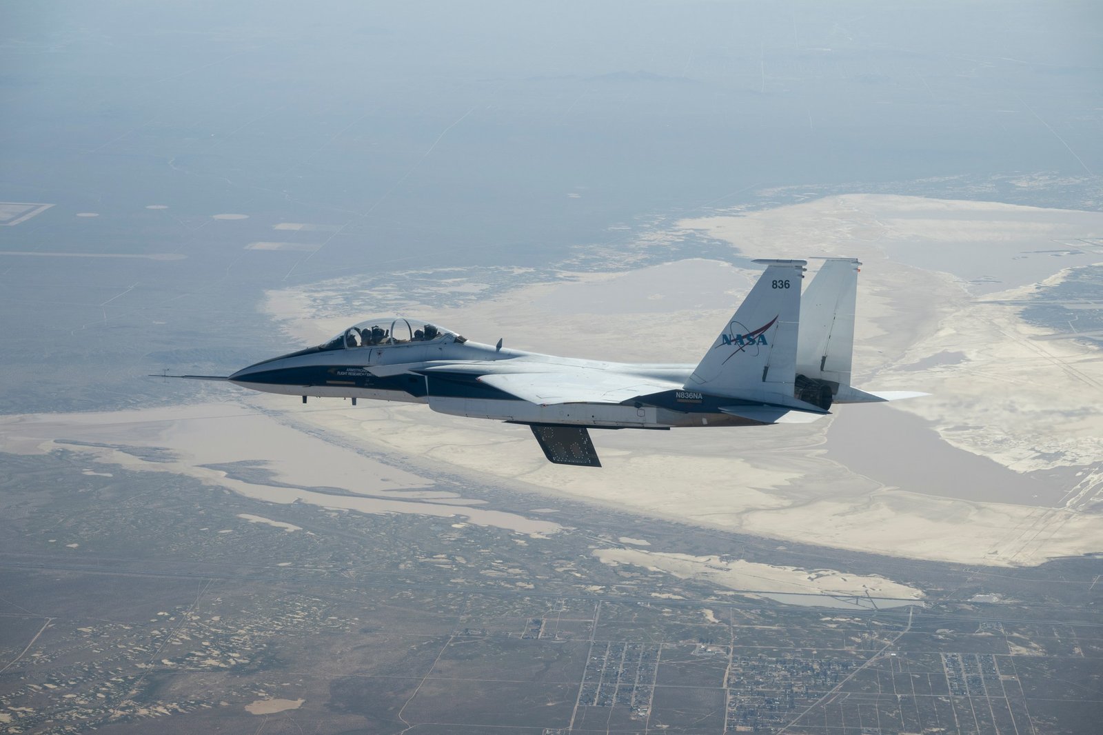 A white and blue NASA F-15 research jet climbs to altitude with an approximately 3-foot experimental wing design mounted beneath its fuselage. Viewed in profile against a blue sky with mountains in the distance, the test article resembles a ventral fin below the aircraft.