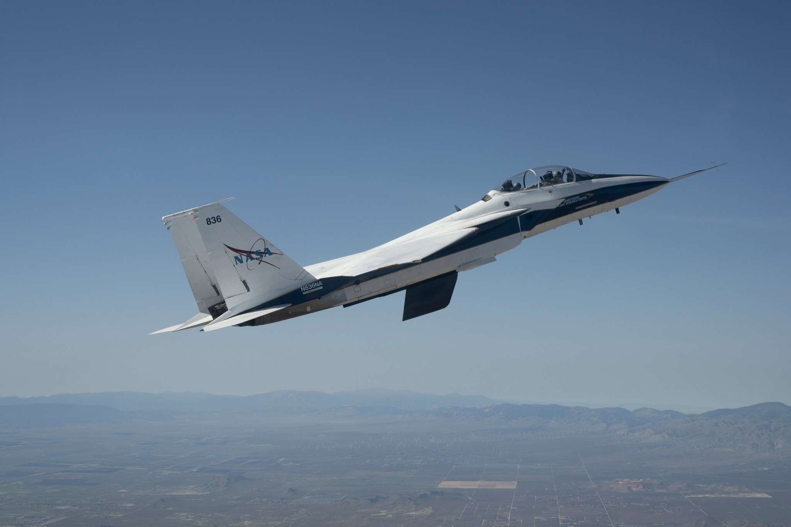 A white and blue NASA F-15 research jet climbs to altitude with an approximately 3-foot experimental wing design mounted beneath its fuselage. Viewed in profile against a blue sky with mountains in the distance, the test article resembles a ventral fin below the aircraft.