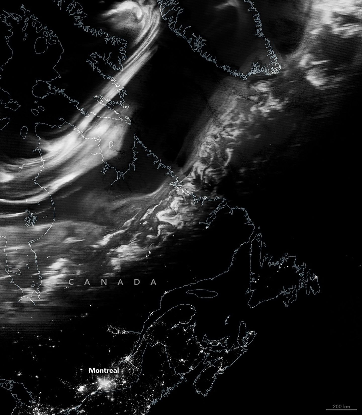 A nighttime satellite image shows a grayscale view of the northern lights stretching from eastern Canada to southern Greenland. Urban light from Montreal and nearby cities appear across the bottom of the scene.