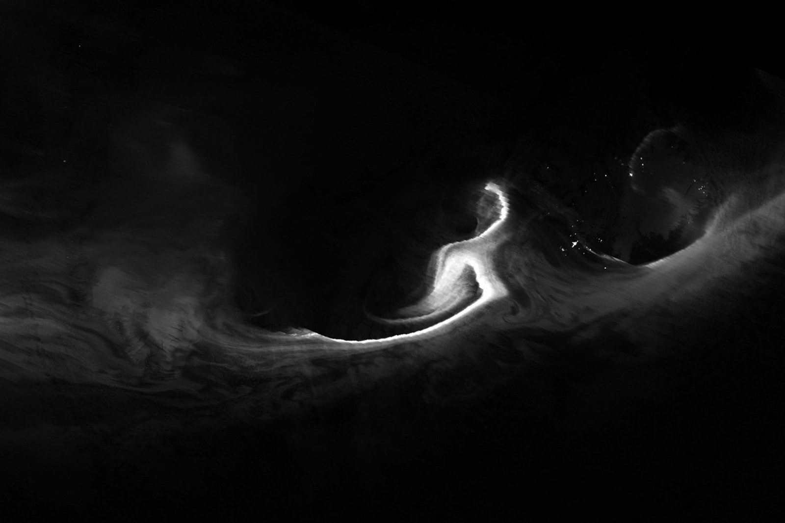 A nighttime satellite image shows a grayscale view of the northern lights over the Denmark Strait. Wisps of light stretch from Greenland to Iceland, with the brightest light just west of Iceland. ReykjavÃk city lights appear as a small dot.