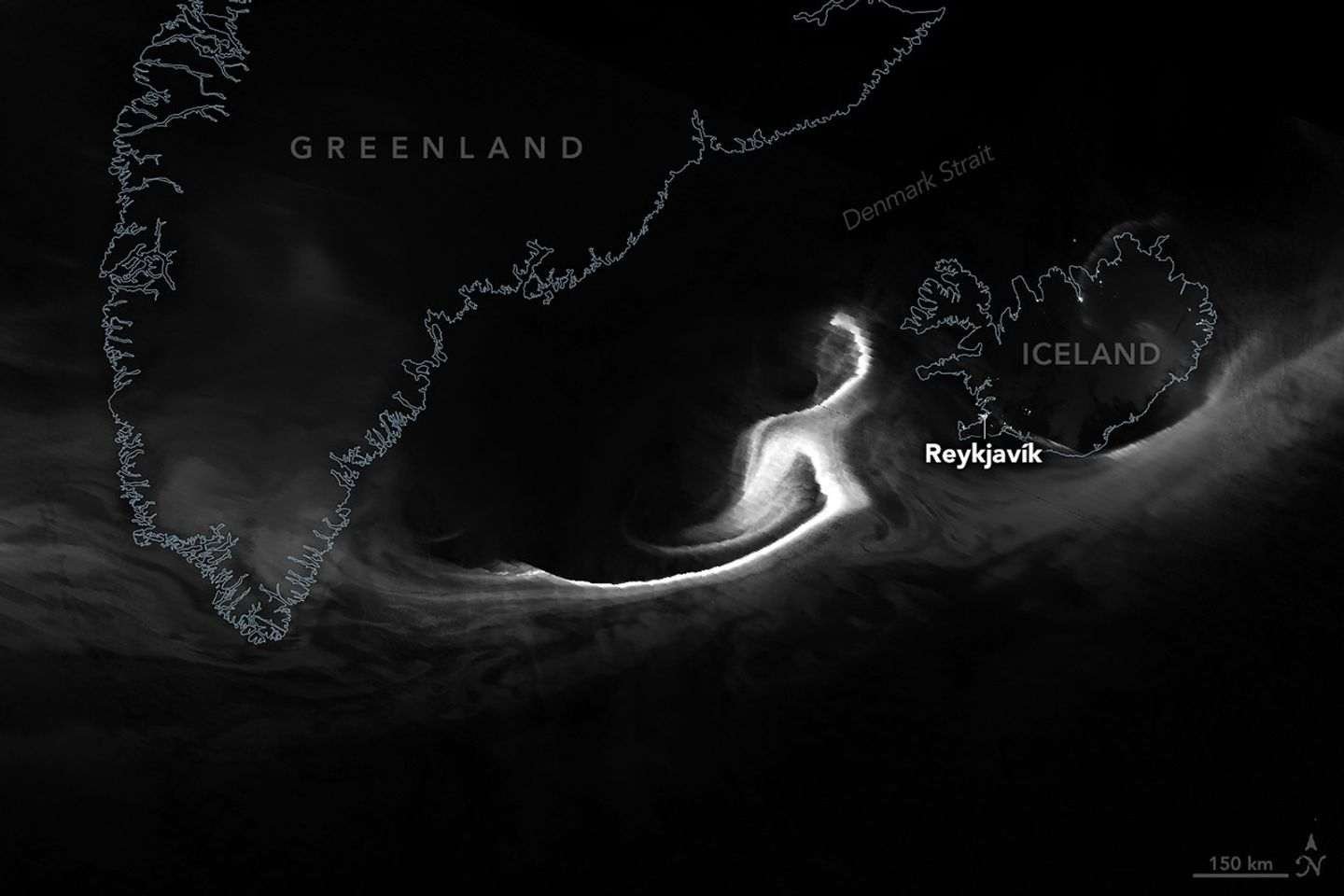 A nighttime satellite image shows a grayscale view of the northern lights over the Denmark Strait. Wisps of light stretch from Greenland to Iceland, with the brightest light just west of Iceland. ReykjavÃk city lights appear as a small dot.