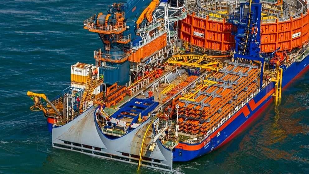 Slightly elevated shot of stern region of flat-bottomed marine vessel laden with a complex and neat assortment of equipment, presumably components of offshore wind systems