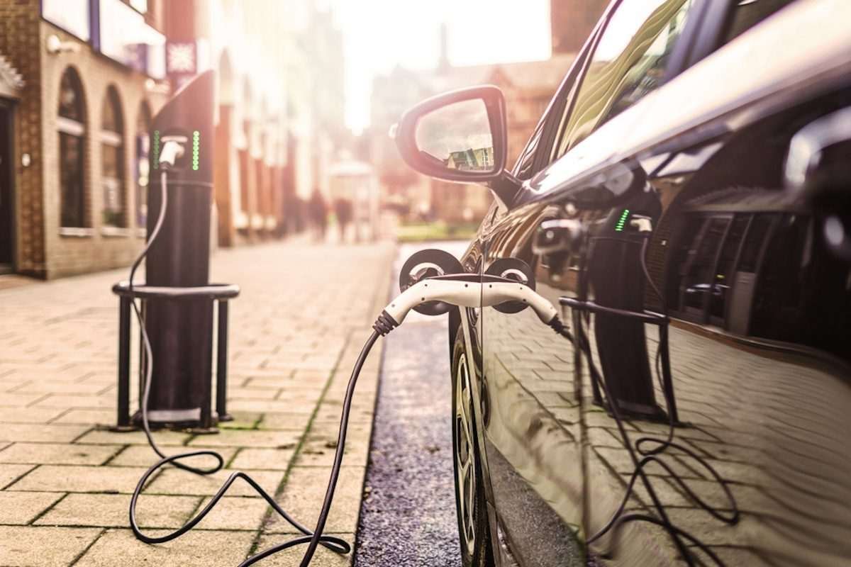 Power inlet on the side of an electric vehicle, with charging underway, and sun-drenched urban scene partially visible in the background