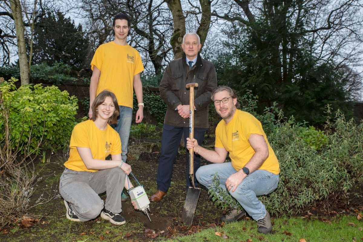 Four people in an environment like a park or garden, one holds a spade, and another holds a cuboidal instrument of some kind from which a long point protrudes into the ground