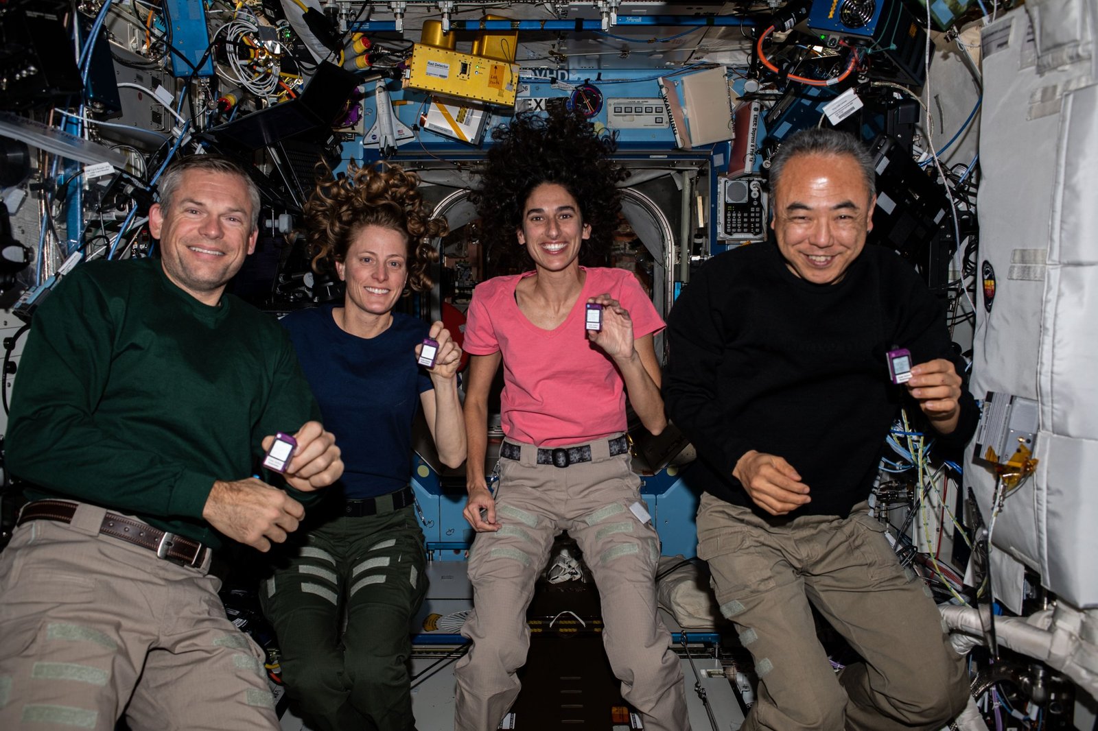 Four astronauts float together inside the International Space Station, smiling and holding small devices about the size of a printer ink cartridge. They are surrounded by equipment, cables, and modules in the microgravity laboratory.
