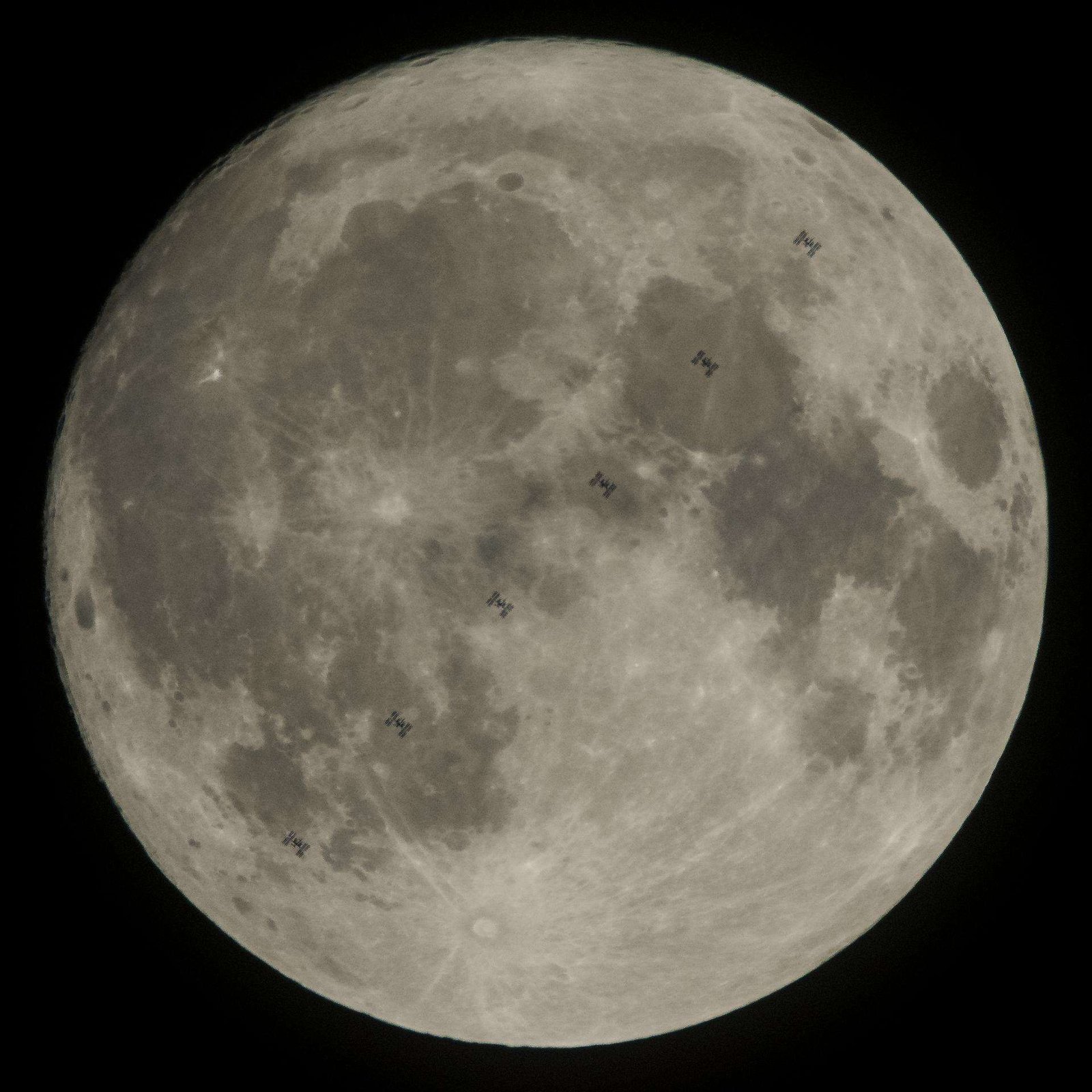 The full Moon fills the frame against a black sky, with multiple small silhouettes of the International Space Station captured as it transits across the Moon’s face.