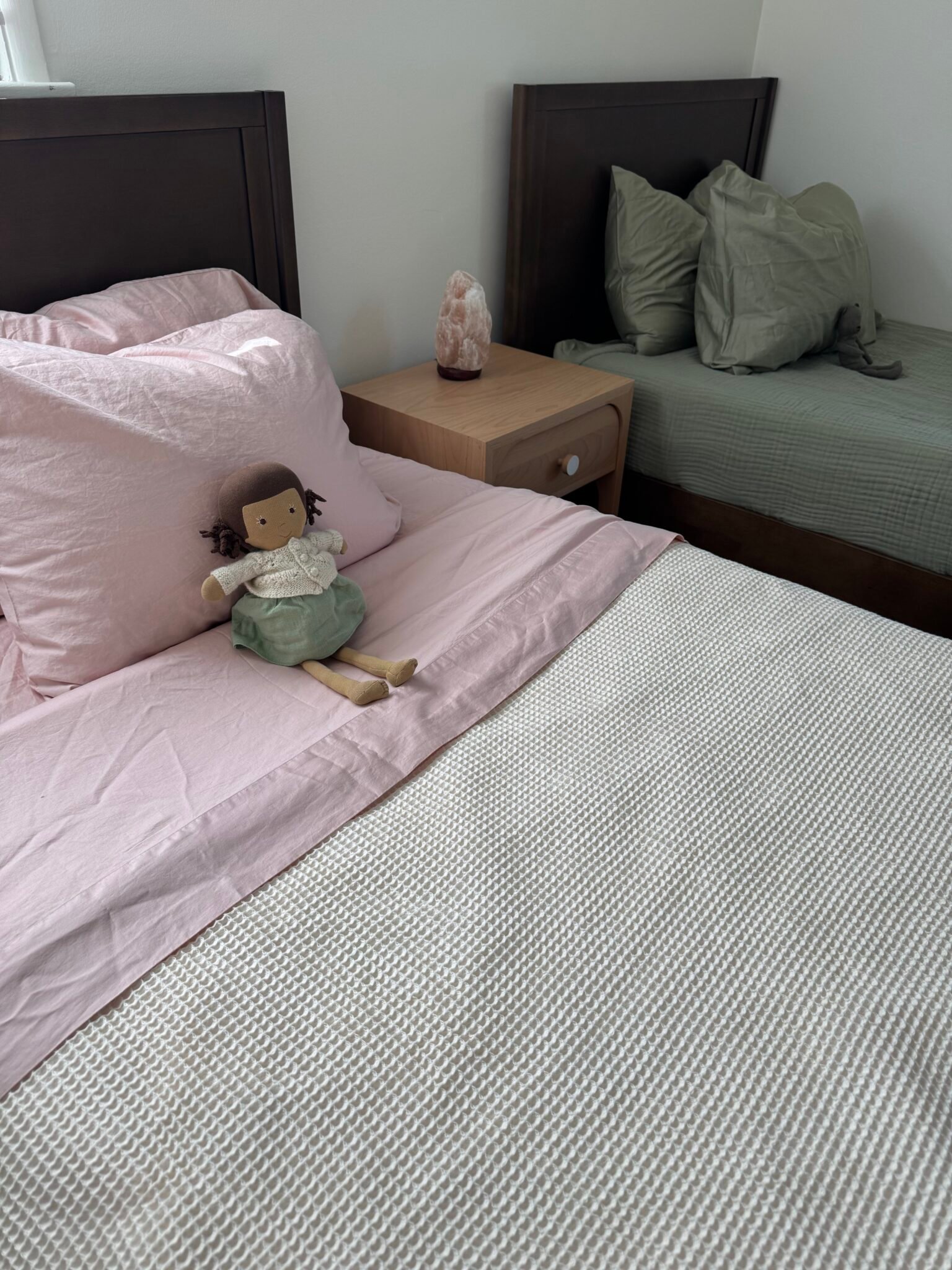 Two neatly made beds with pastel bedding, a nightstand between them, and a cloth doll placed on the bed with pink sheets.