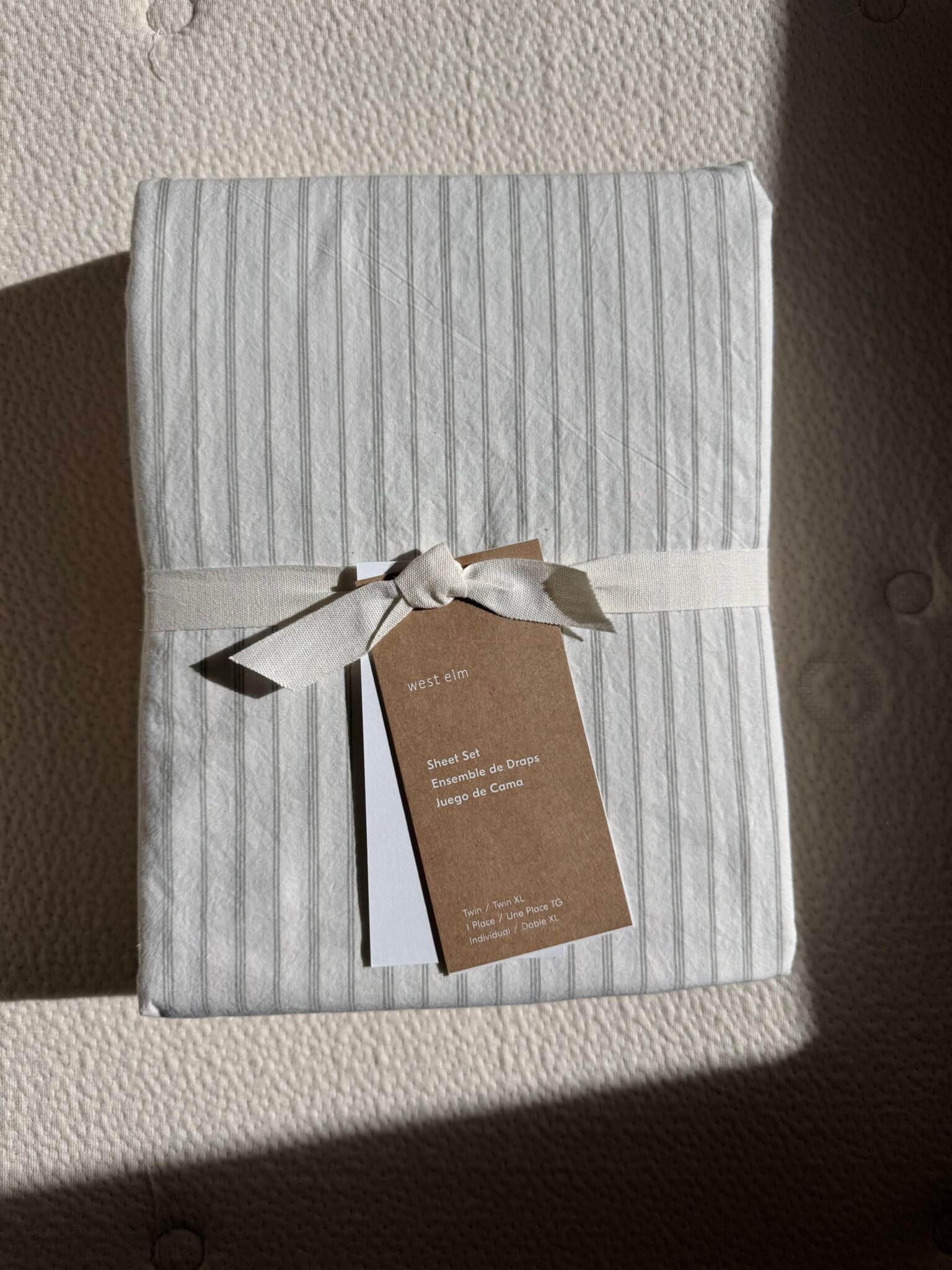 A neatly wrapped set of striped sheets with a cream-colored ribbon and a brown tag, placed on a textured surface in sunlight.