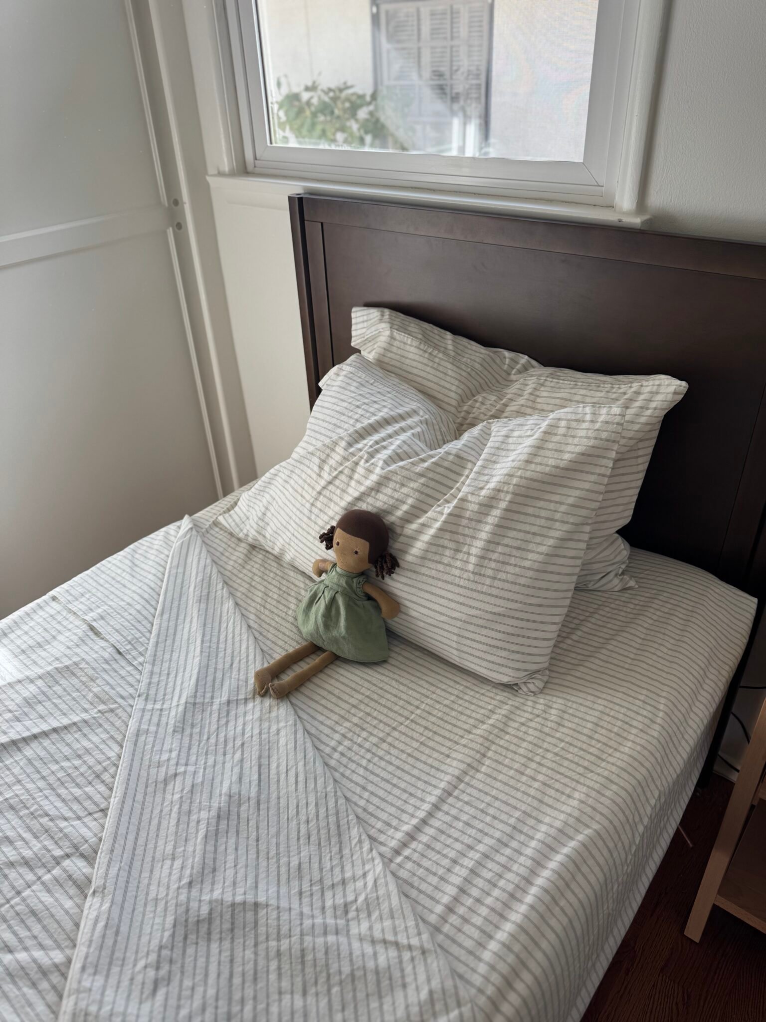 A neatly made single bed with striped white bedding and a fabric doll in a green dress placed on the pillow, beside a window with natural light.