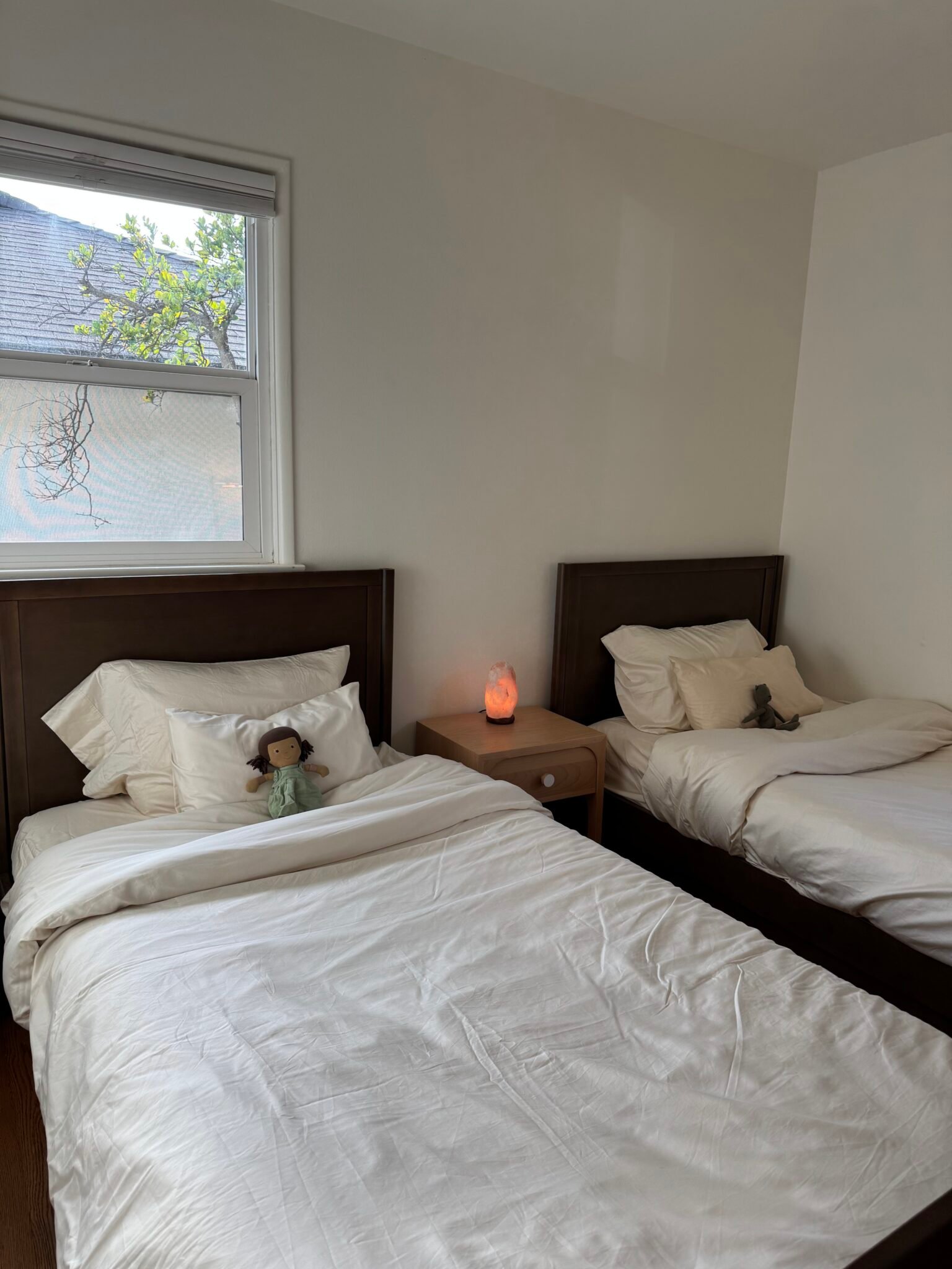 Two single beds with white bedding are placed in a corner of a room. A nightstand with a salt lamp sits between them. Each bed has a doll or stuffed animal on the pillow.