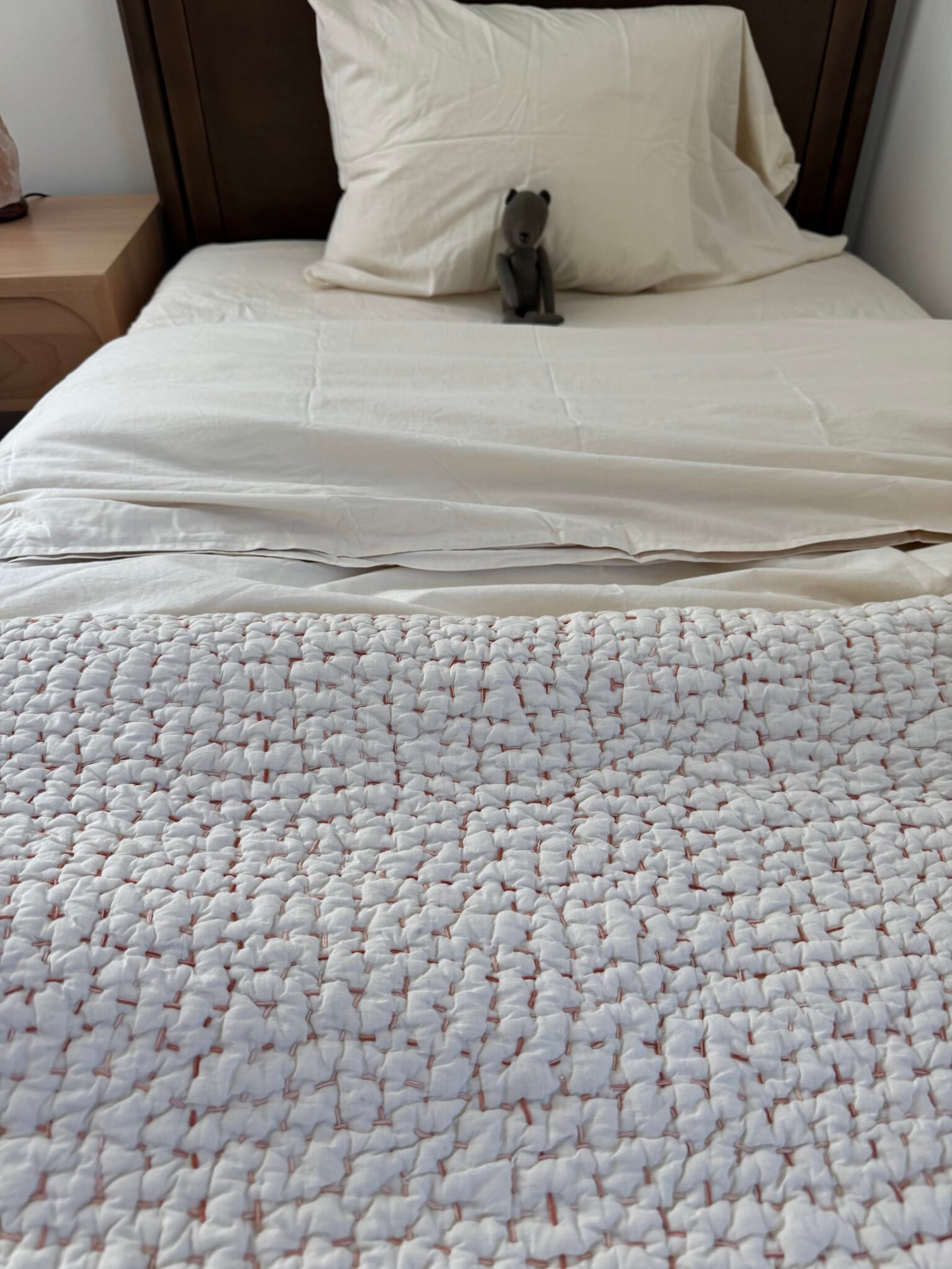 A neatly made bed with white bedding, a textured white blanket at the foot, and a small stuffed animal placed against the pillow.