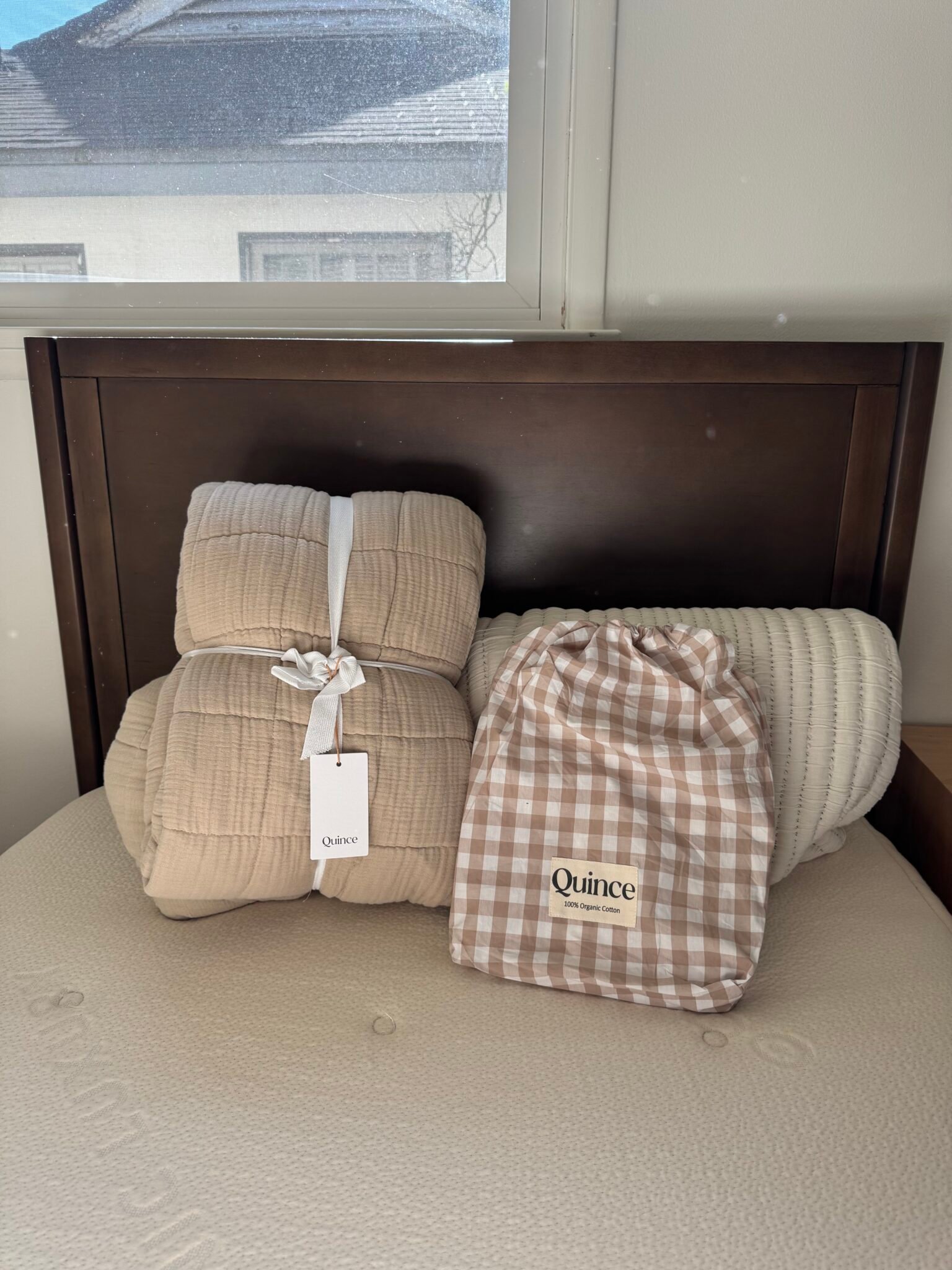 A beige quilt, a white bolster pillow, and a gingham Quince bag are placed on a mattress against a dark wood headboard under a window.