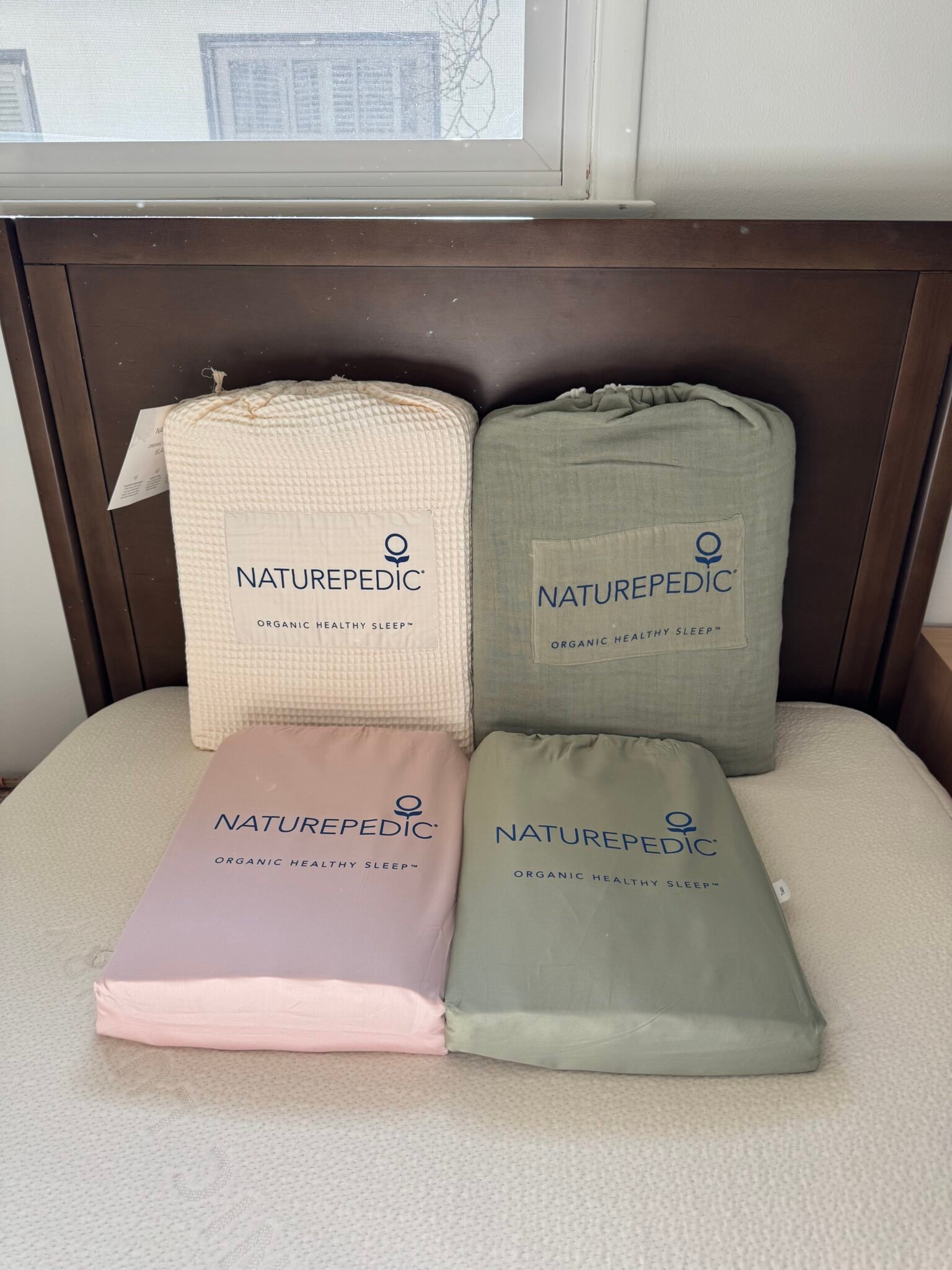 Four packaged Naturepedic bedding items in assorted colors (cream, green, pink, and sage) are displayed upright on a bed with a dark wooden headboard.