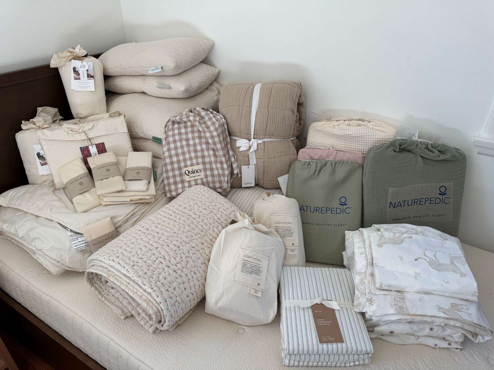 A variety of neatly stacked bedding items, including pillows, quilts, comforters, sheets, and blankets, are arranged on a bed against a white wall.