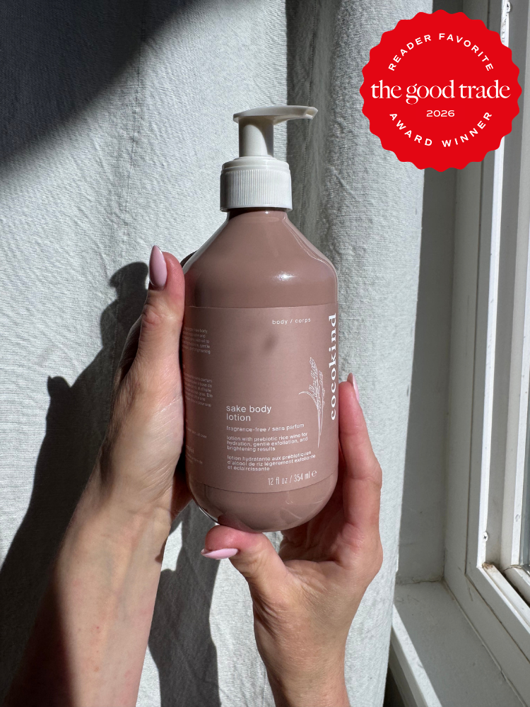 A person holds a bottle of Cocokind sake body lotion near a sunlit window. A red badge in the corner reads "The Good Trade Award Winner 2026 Reader Favorite.