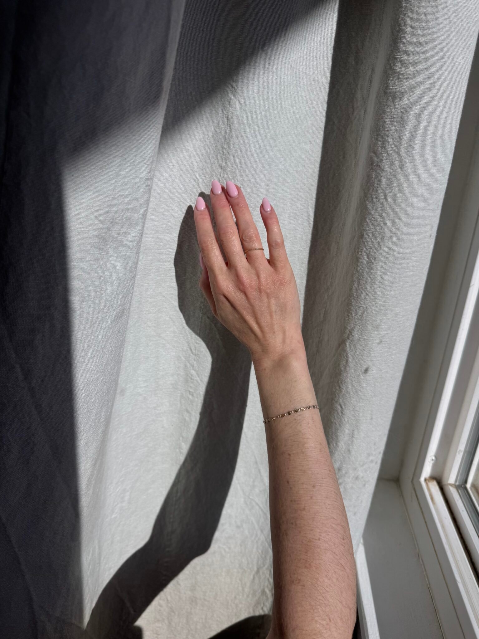 A hand with light pink nails touches a sunlit white curtain, casting a shadow on the fabric near a window.