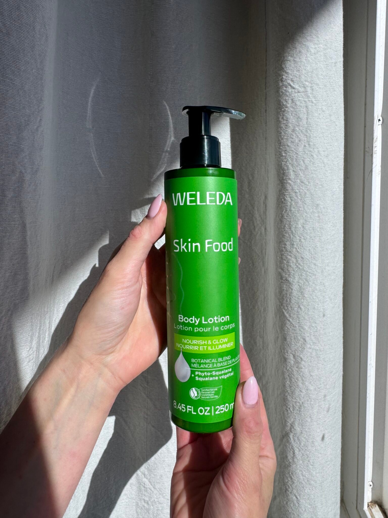A person holds a green bottle of Weleda Skin Food Body Lotion, 8.45 fl oz, with a pump dispenser, in natural light by a white curtain.