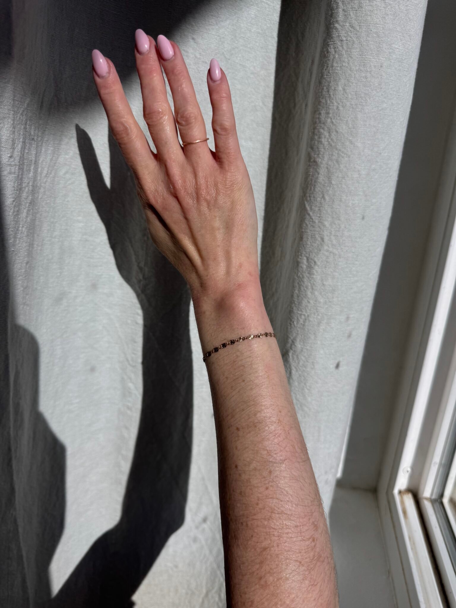 A hand with light pink nail polish and a thin bracelet is raised against a sunlit white curtain, casting a shadow.