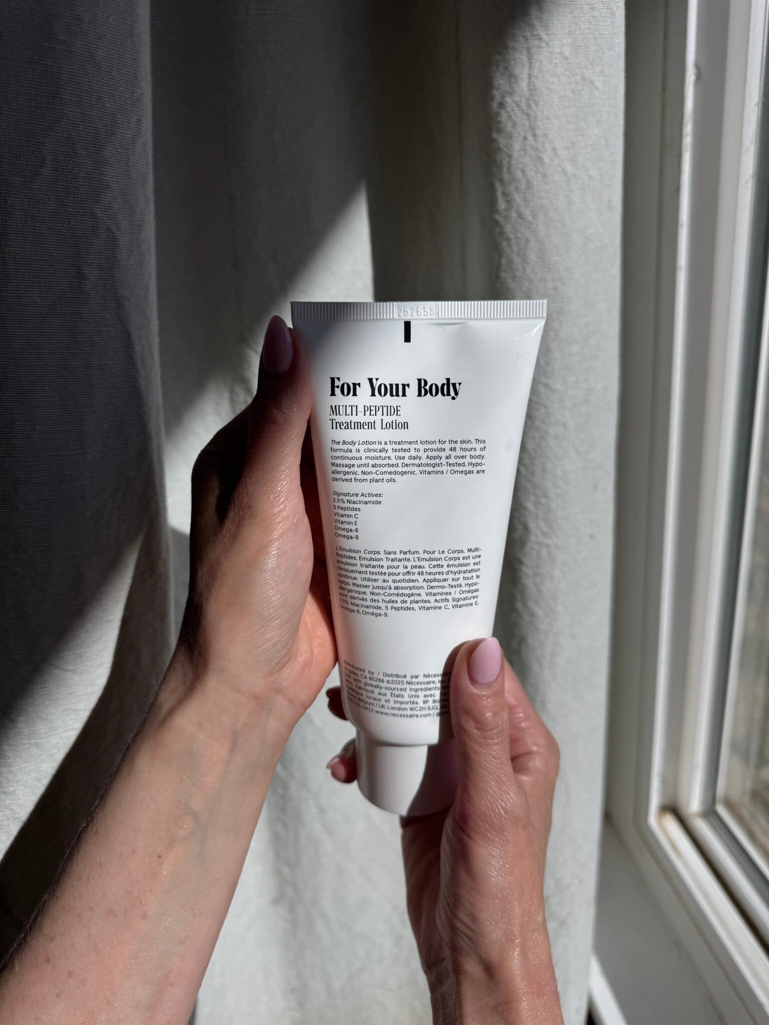 Person holding a white tube labeled "For Your Body Multi-Peptide Treatment Lotion" near a sunlit window, with the text on the tube partially visible.
