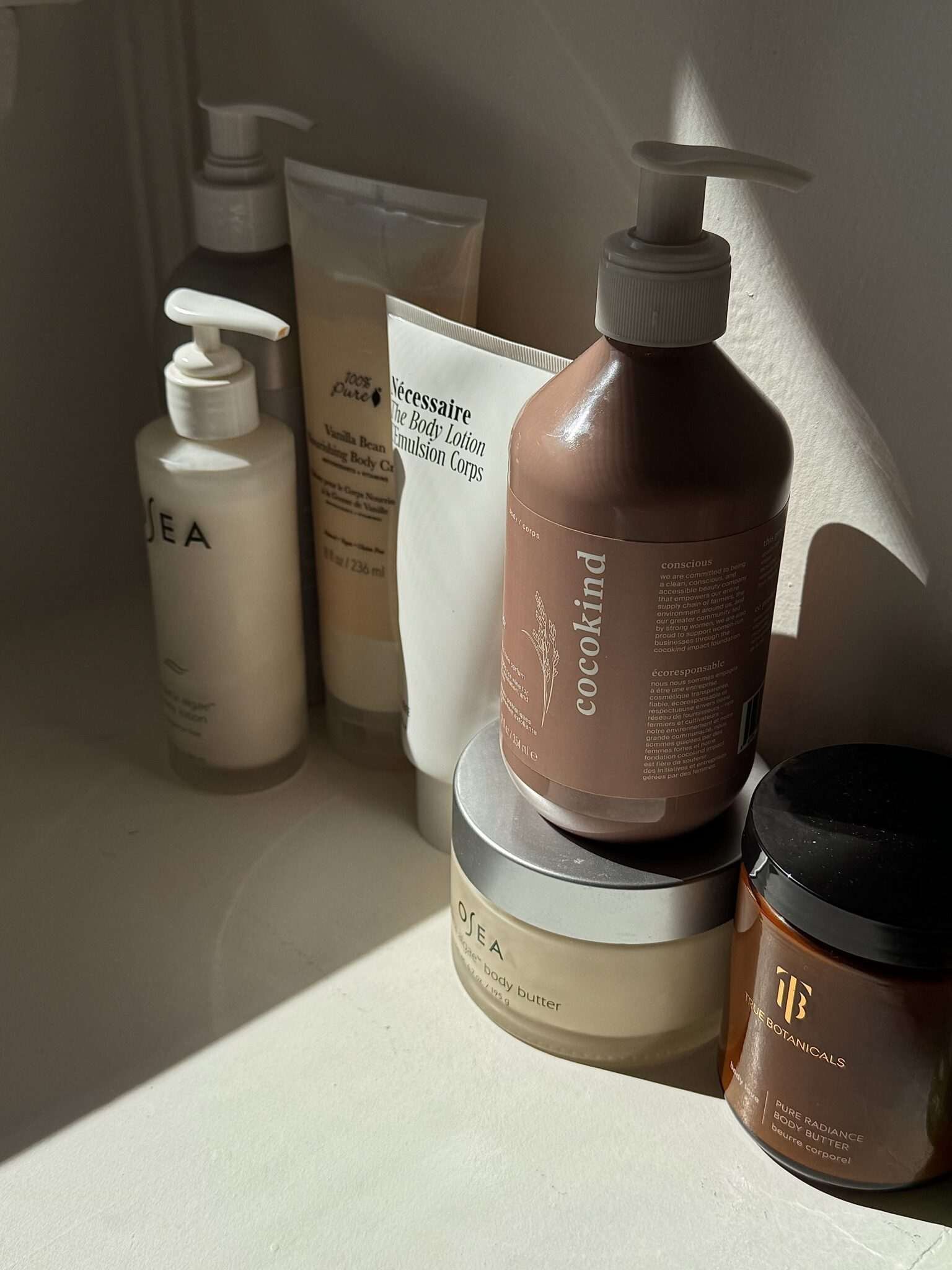 A collection of skincare and body care products, including lotions, creams, and a jar, arranged on a white surface in natural light.