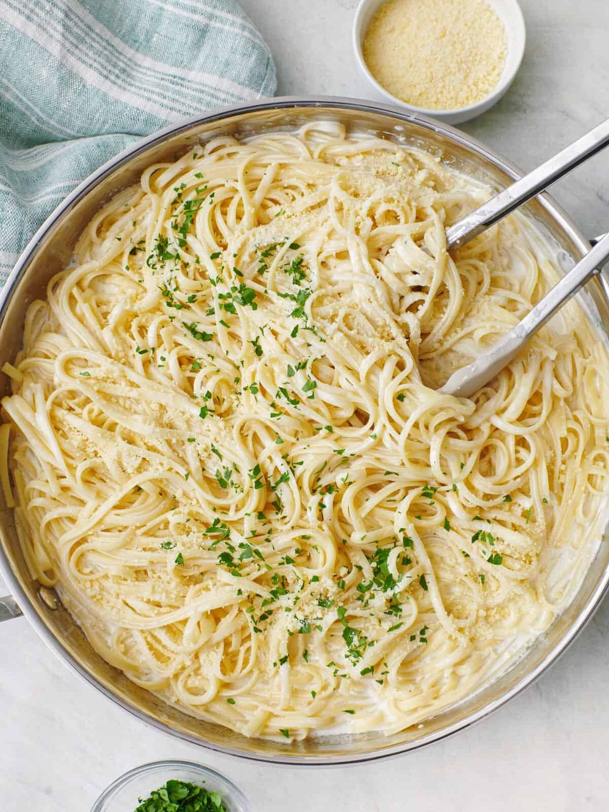 A pan of fettuccine Alfredo topped with chopped parsley and grated cheese, with metal tongs, on a marble surface next to small bowls of cheese and parsley.