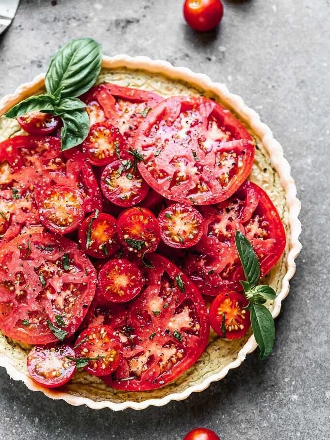 A tomato tart topped with sliced tomatoes, cherry tomatoes, fresh basil leaves, and black pepper on a gray surface.