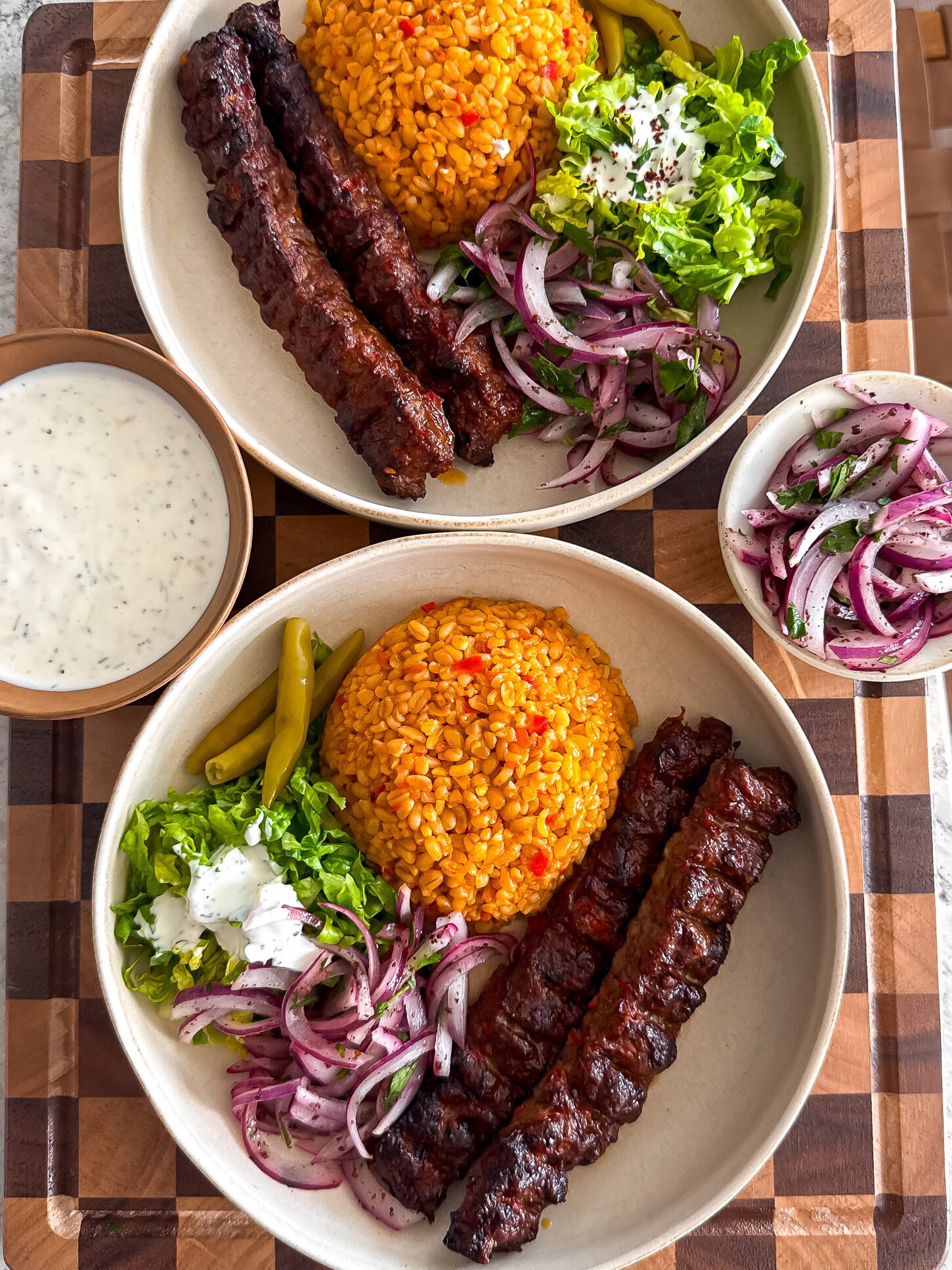 Two plates with grilled kebab skewers, spiced rice, sliced red onions, lettuce, green peppers, and yogurt sauce on a wooden board.