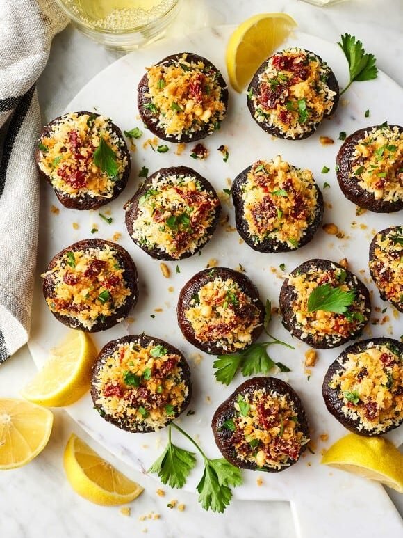 A marble platter with stuffed mushrooms topped with breadcrumbs and herbs, garnished with parsley and lemon wedges on the side.