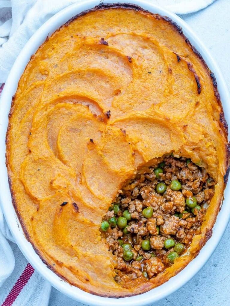 Oval dish of shepherd's pie with a section removed, showing ground meat, peas, and sauce under a layer of browned mashed potatoes.