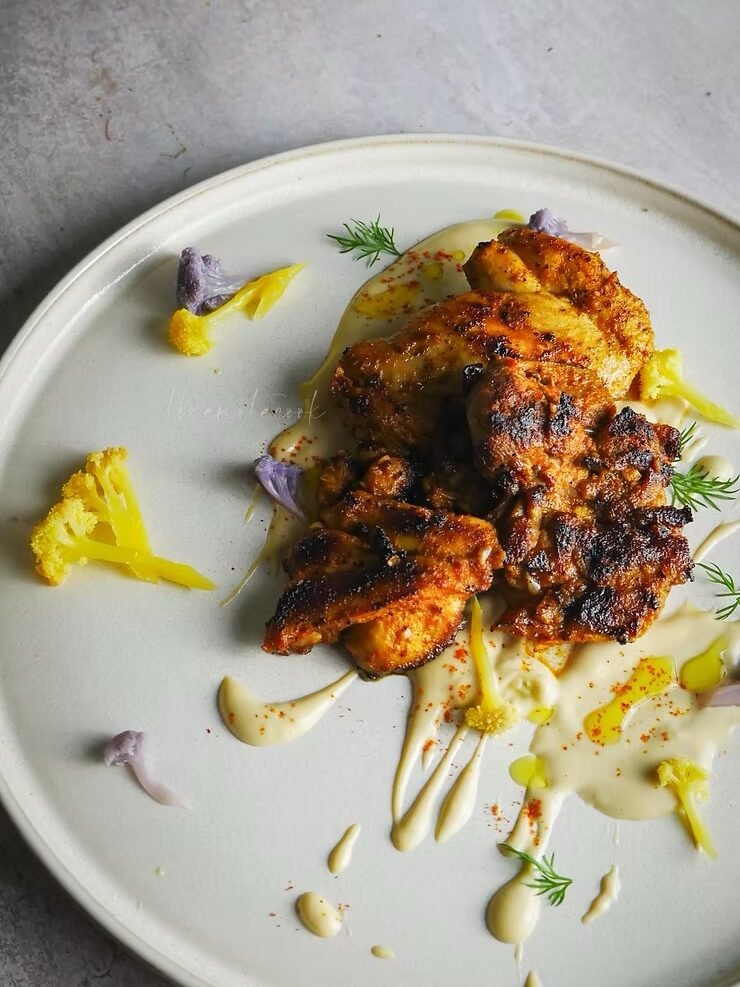 Grilled chicken pieces on a white plate with creamy sauce, garnished with small florets of yellow and purple cauliflower and fresh herbs.