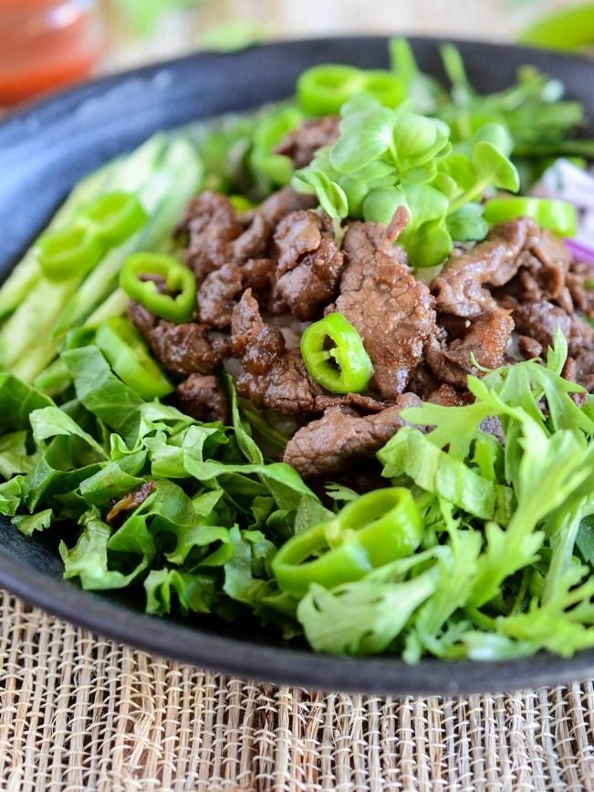 A black bowl filled with sliced beef, green lettuce, and sliced green chili peppers on a woven placemat, with a small bowl of red sauce in the background.
