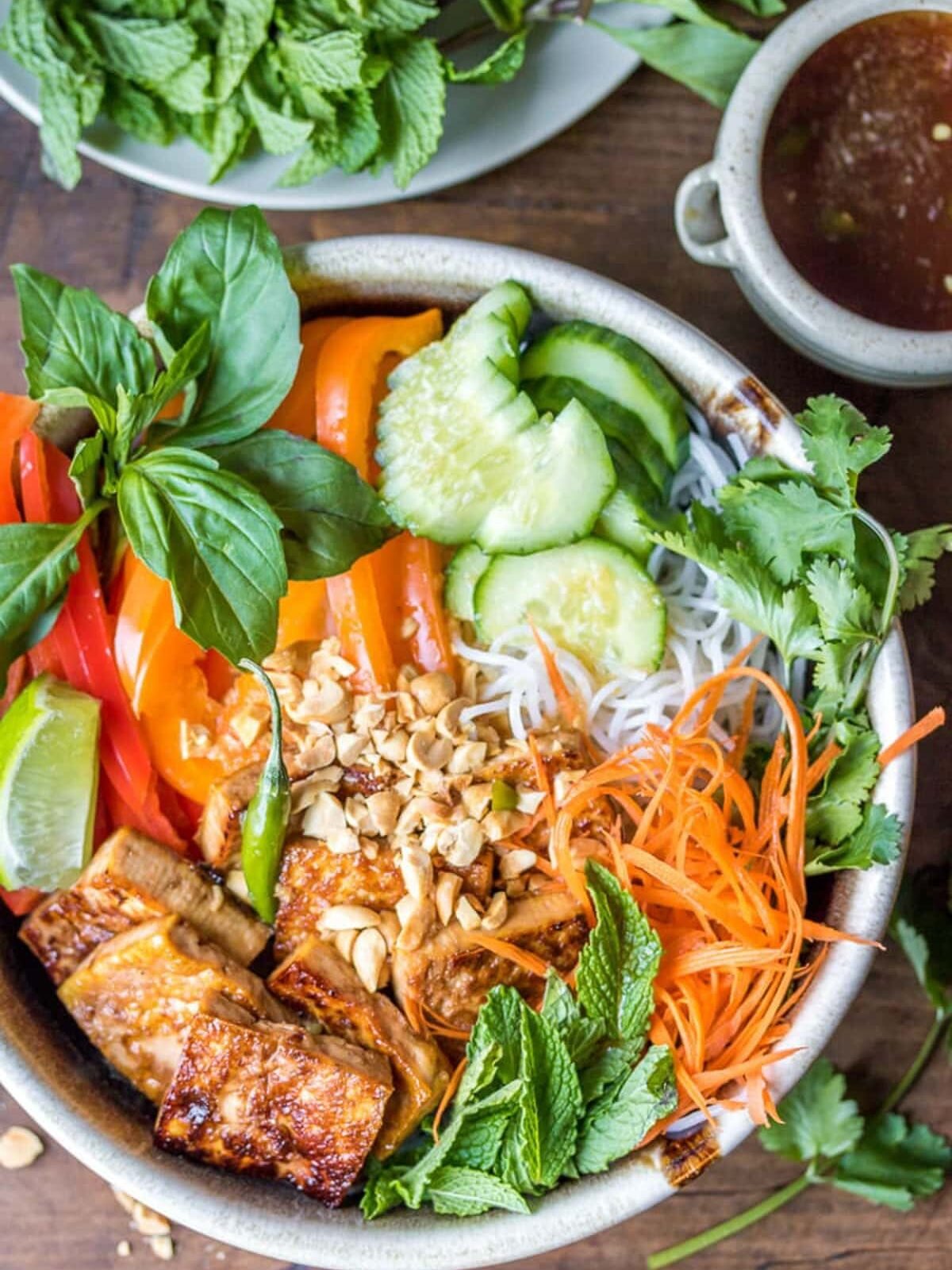 A bowl of Vietnamese vermicelli noodles with grilled tofu, sliced bell peppers, cucumber, carrots, mint, basil, cilantro, lime wedge, and chopped peanuts, with sauce on the side.