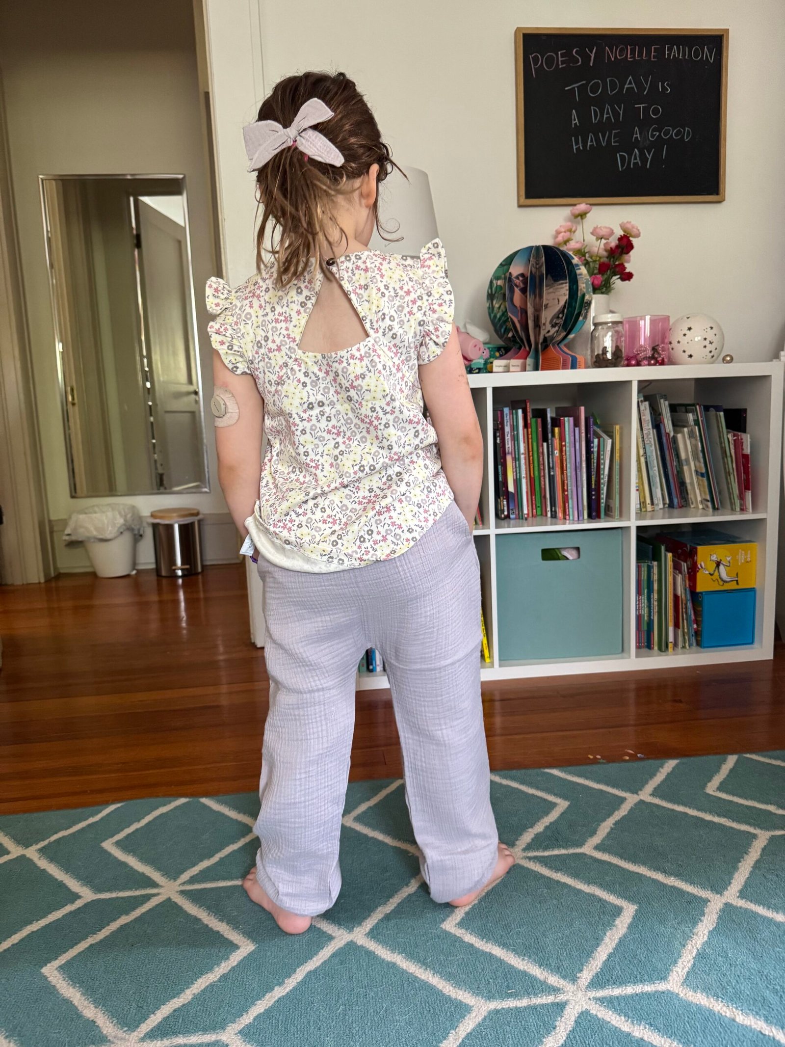 A young child with a bow in their hair stands barefoot on a blue rug, facing away, in a room with a bookshelf, a mirror, and a chalkboard with writing.