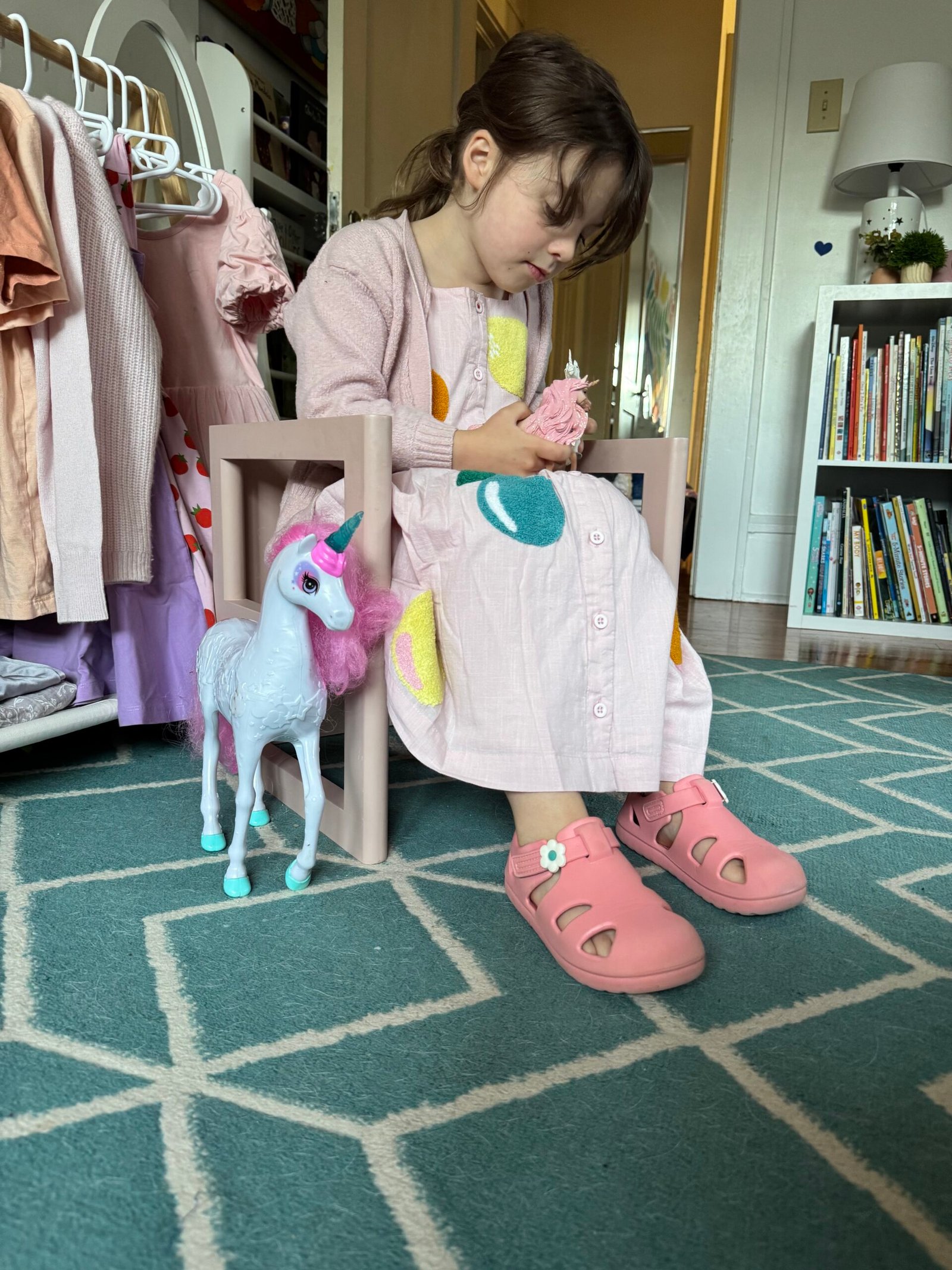 A young girl in a pink dress and shoes sits on a small chair holding a toy, with a unicorn figurine on the floor beside her in a room with bookshelves and clothes.