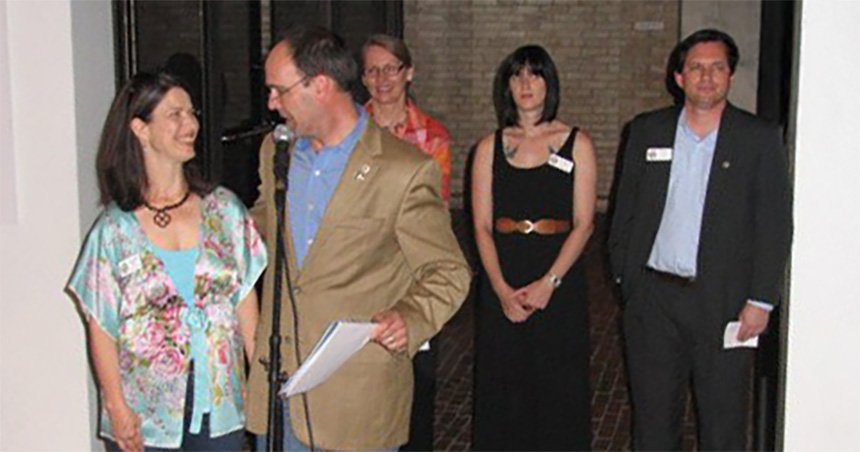 A group of six adults stands together in an indoor event space with brick walls and a tiled floor. A man in a tan blazer speaks into a microphone while holding papers, and a woman in a floral blouse smiles beside him. Four other people stand behind them wearing business or event attire with name tags, suggesting a presentation or announcement.