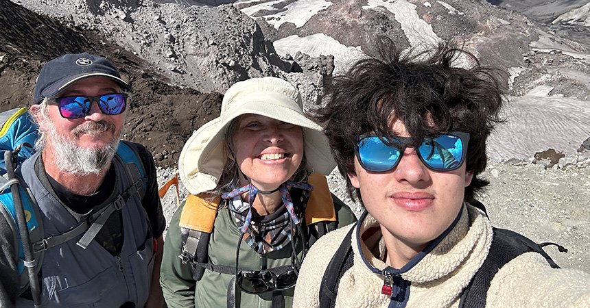 Three hikers take a close-up selfie on a rocky, high?altitude mountain slope. All three wear outdoor gear—backpacks, hats, and sunglasses—and smile toward the camera. Behind them, steep gray rock and patches of snow or glacier rise sharply under bright sunlight, suggesting a rugged alpine environment.