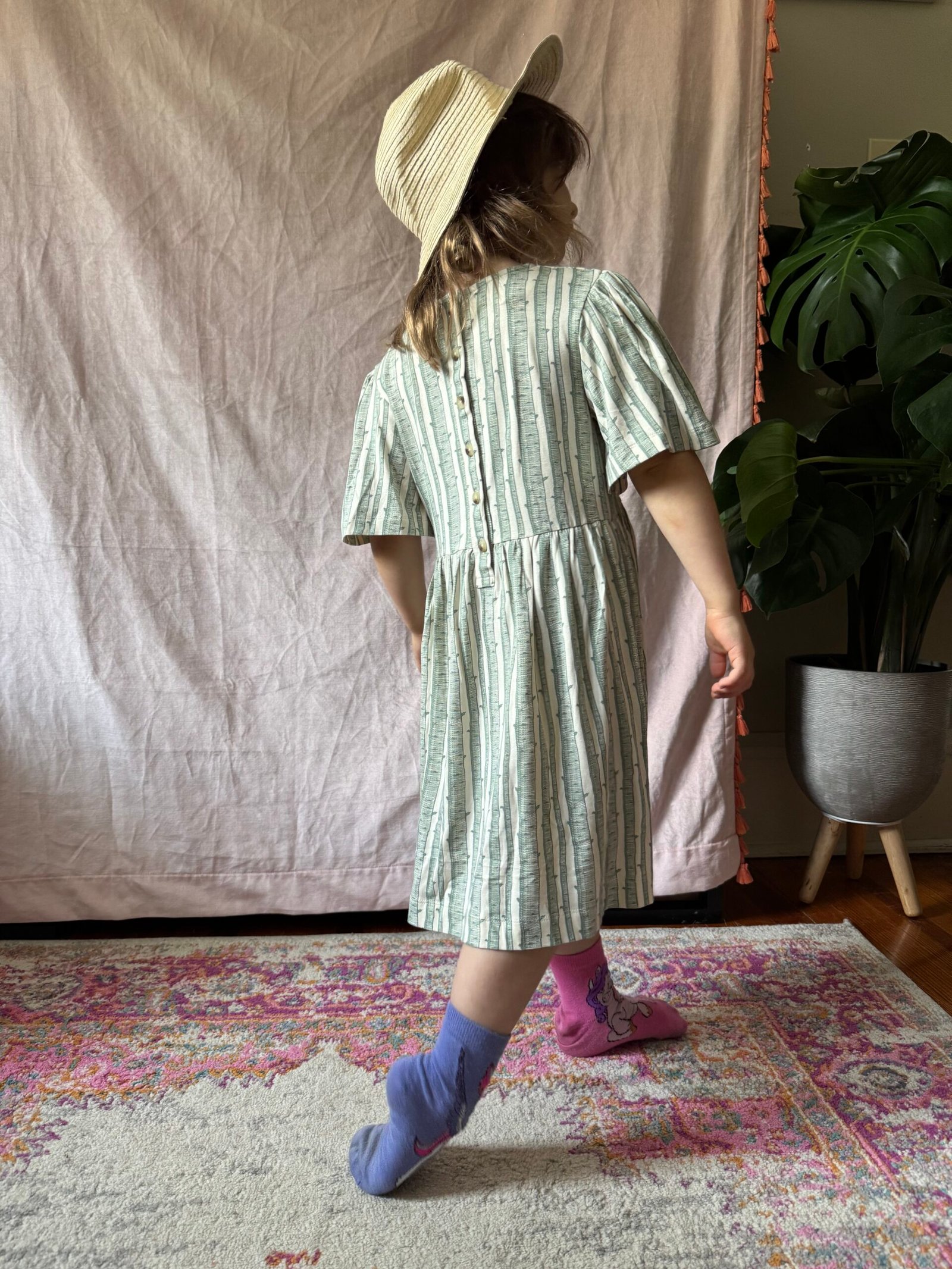 A child wearing a striped dress, hat, and mismatched socks stands on a patterned rug with their back turned, near a large green potted plant and a light pink fabric backdrop.