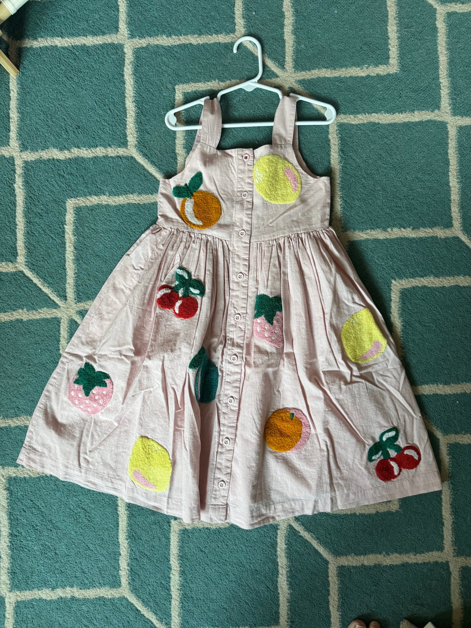 A pink sleeveless dress with button-down front, decorated with large embroidered fruit designs, displayed on a hanger over a geometric patterned rug.