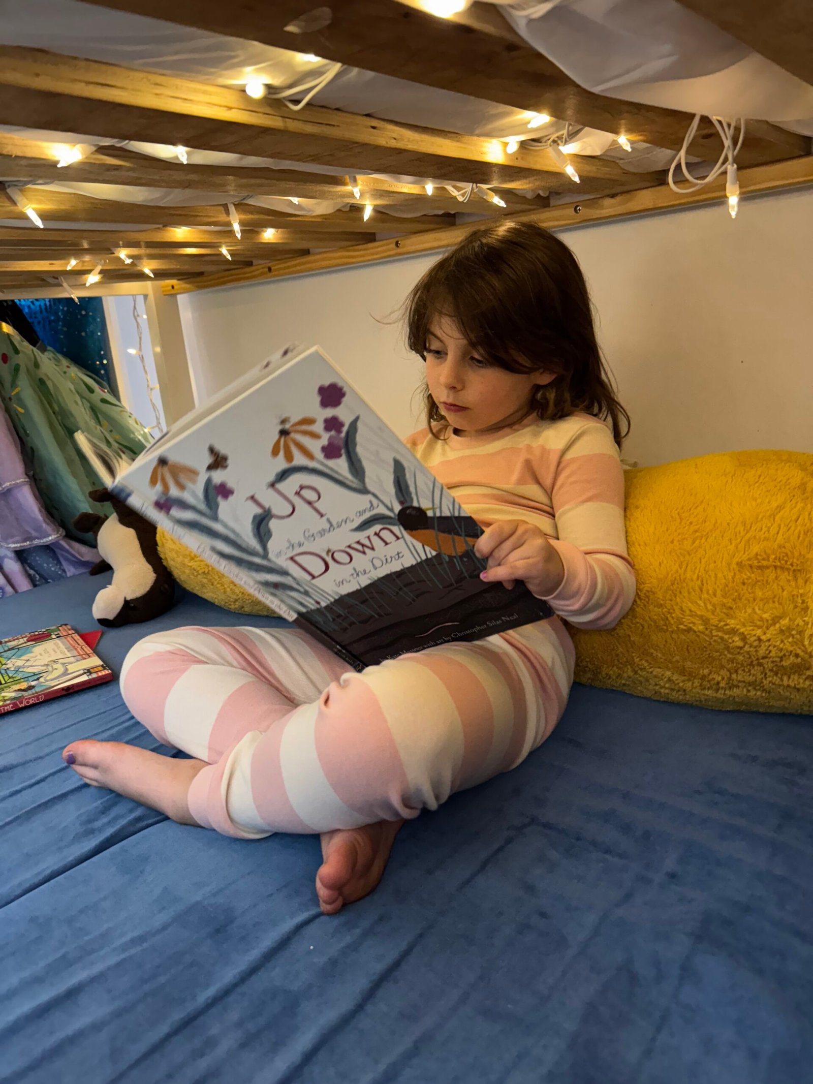 A young child in striped pajamas sits on a blue bed with a yellow pillow, reading a picture book under warm string lights.