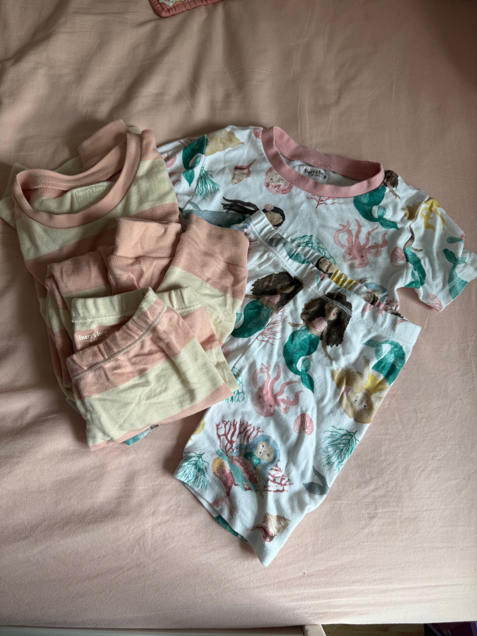 Two sets of children's pajamas are laid out on a pink bedspread; one set has pink and cream stripes, the other features a mermaid and ocean-themed print with pink trim.