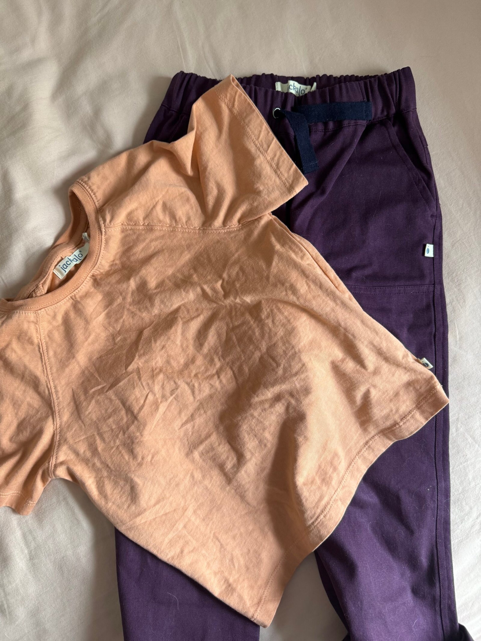 A peach-colored short-sleeve t-shirt is laid on top of a pair of dark purple pants on a beige surface.