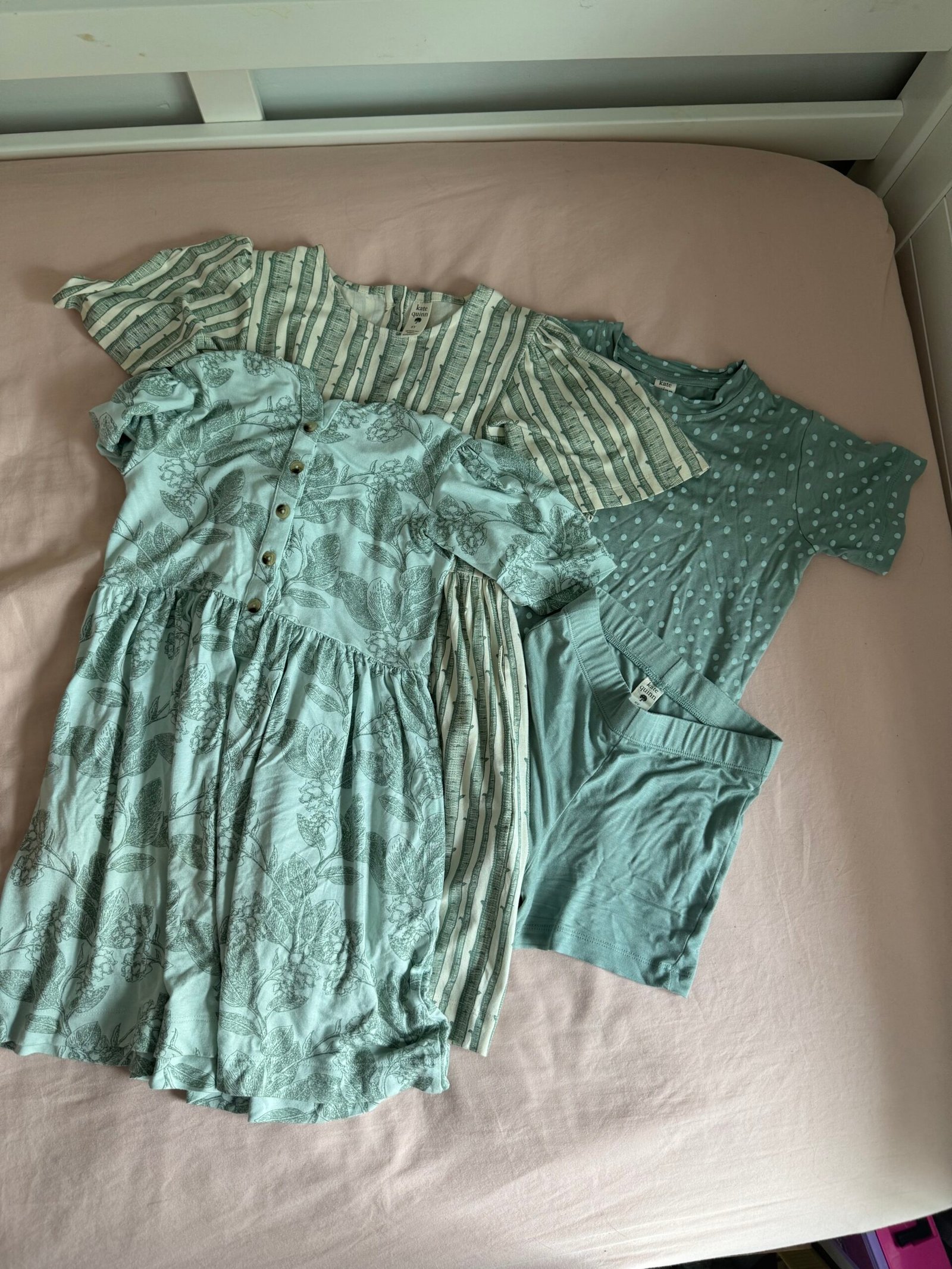 Four pieces of children’s clothing in shades of green are laid out on a pink bedsheet, including two dresses, a t-shirt, and a pair of shorts.