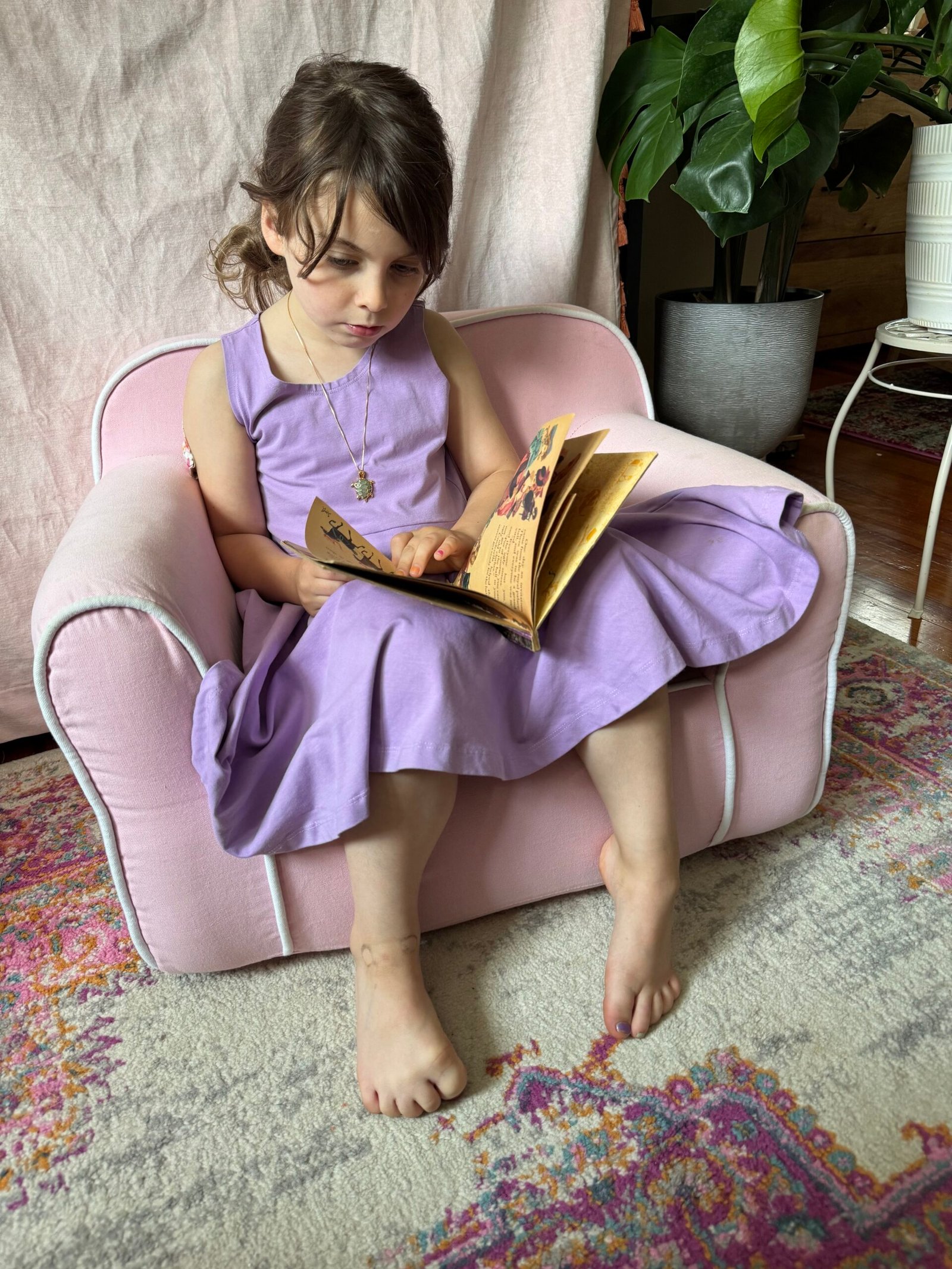 A young girl in a purple dress sits barefoot on a pink chair, reading a book. There is a plant and a patterned rug nearby.
