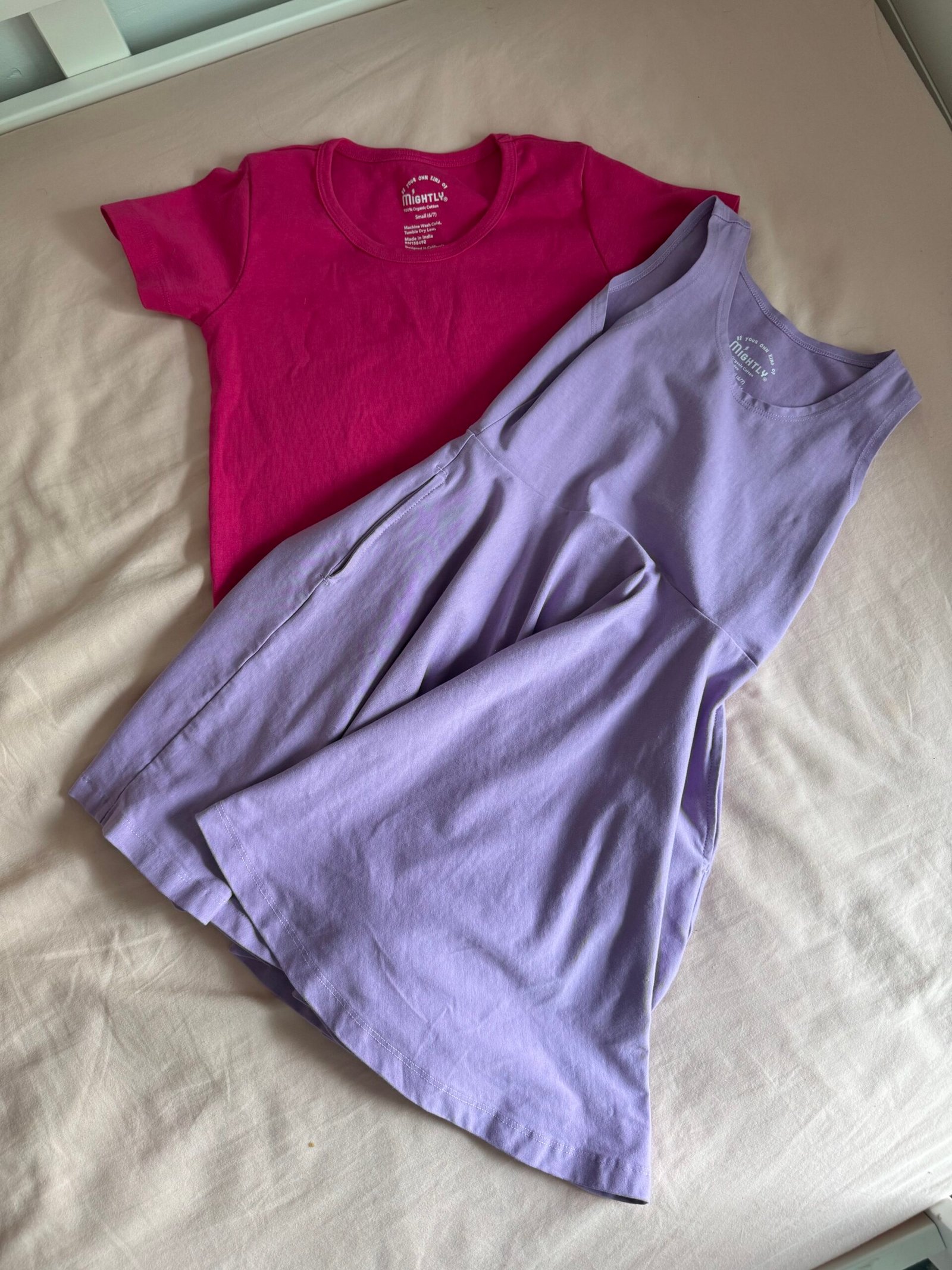 A pink short-sleeve t-shirt and a sleeveless lavender dress are laid out on a light beige surface.