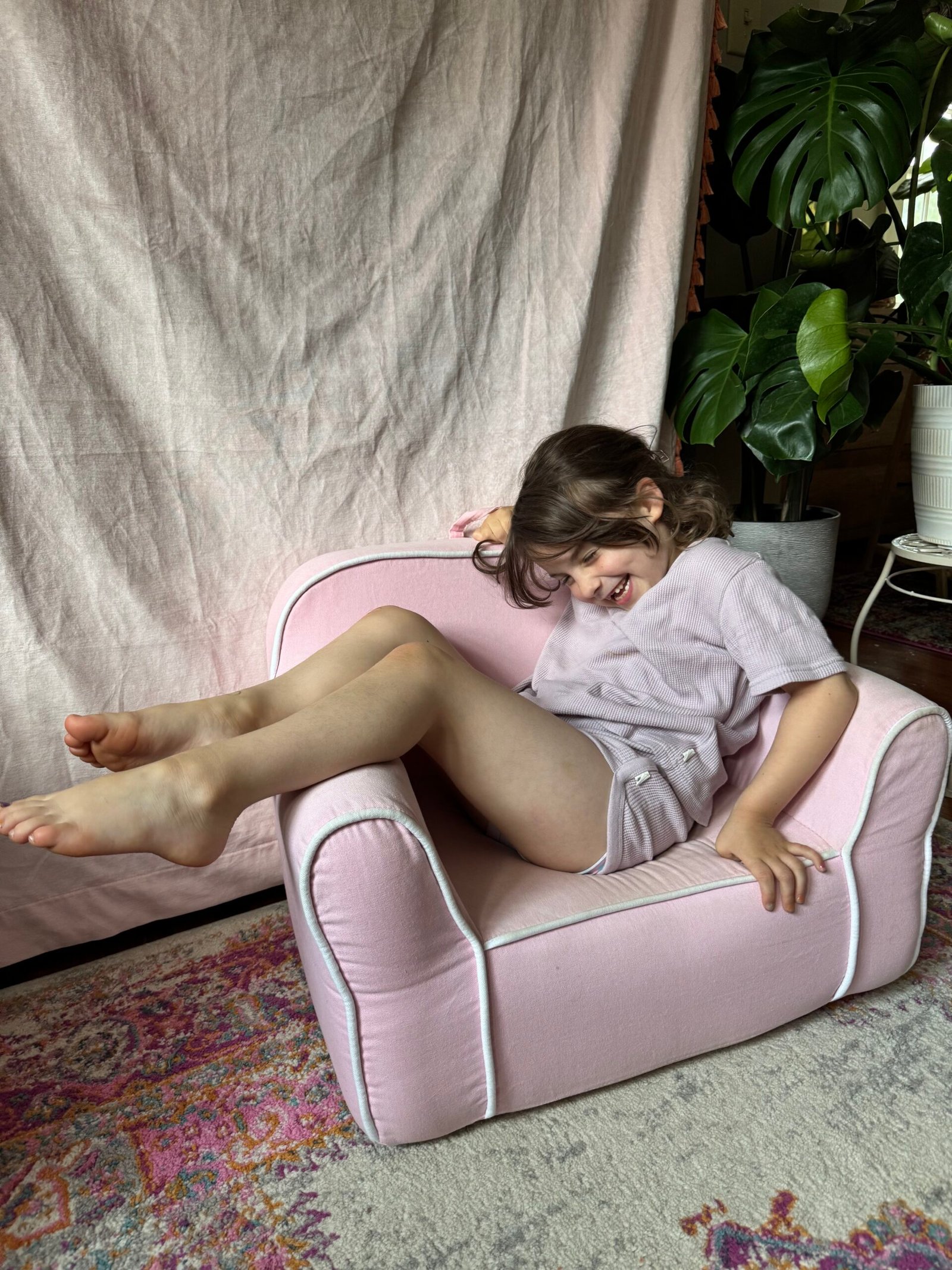 A young child sits sideways and smiles on a small pink armchair, with her legs resting over one arm. A plant and a pink fabric backdrop are visible in the background.