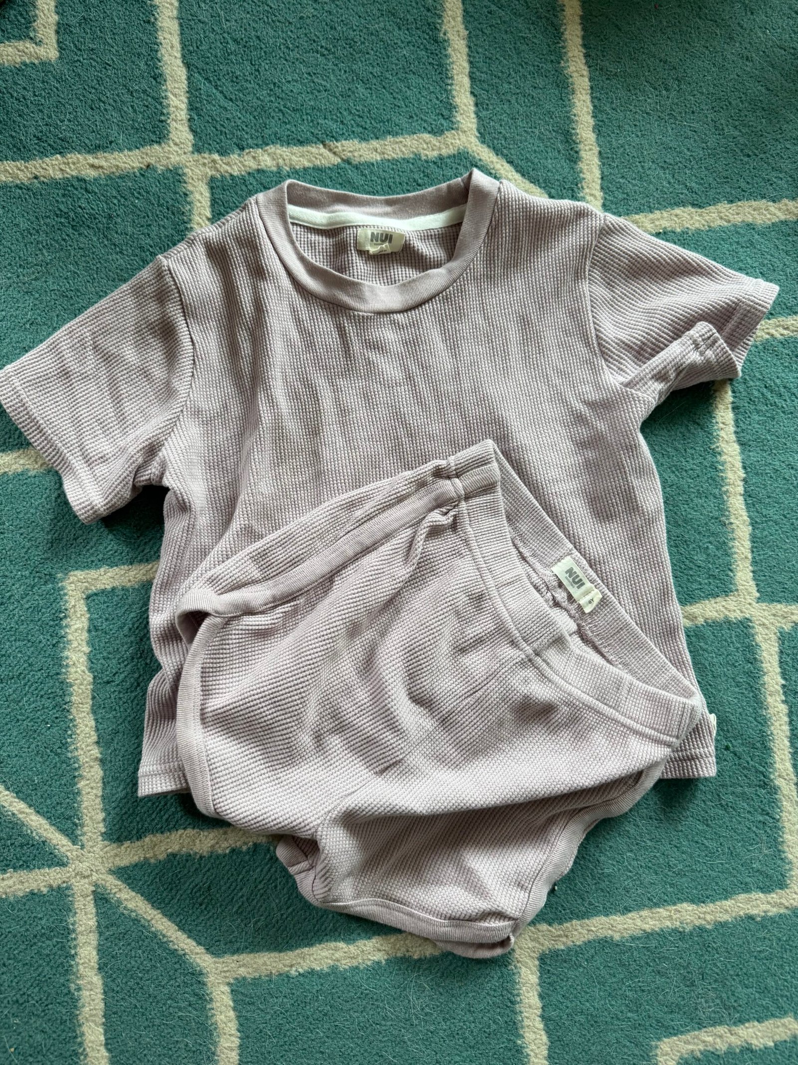 A short-sleeve ribbed lavender top and matching shorts set are laid out on a blue and white geometric-patterned rug.
