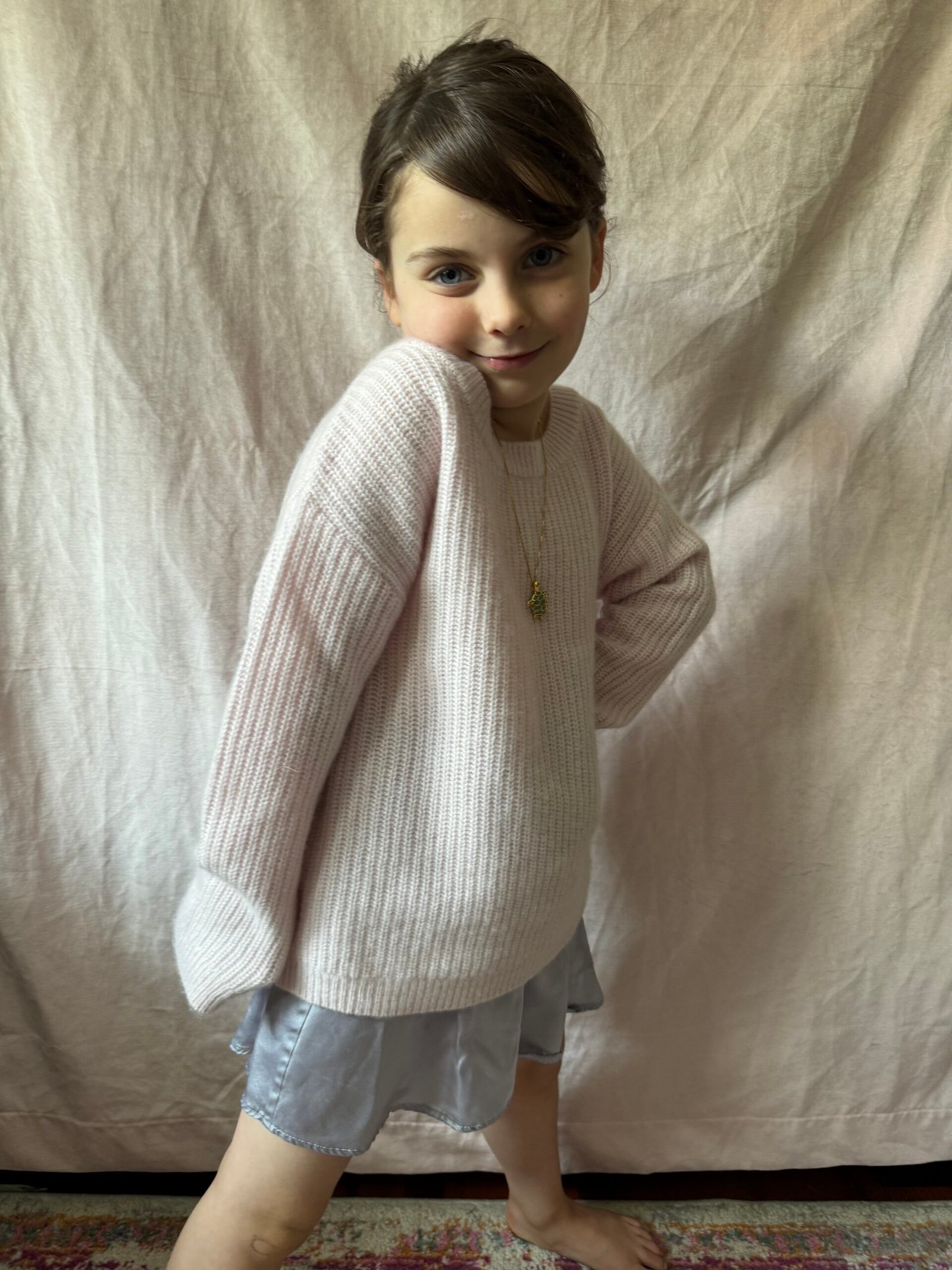 A young child stands in front of a light-colored fabric backdrop, wearing a pale pink sweater, a gray skirt, and a necklace, with one hand on their hip and a slight smile.