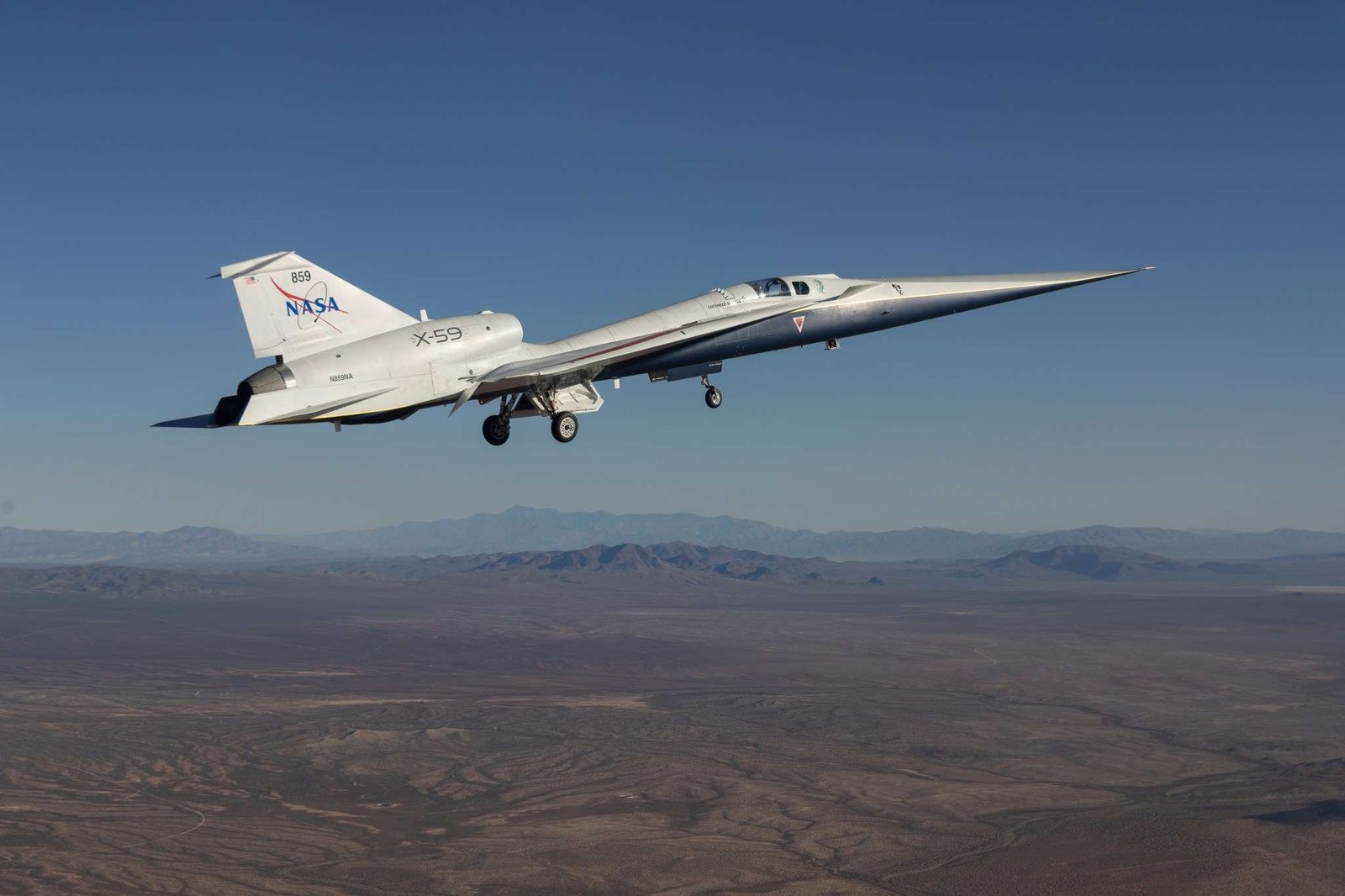 Image of a sleek, white airplane with a sharp, pointed nose flies above arid mountains. The plane's wheels are down. NASA is painted in blue lettering on its tail. The X-59 has a role in NASA's Flight Demonstrations and Capabilities (FDC) project.